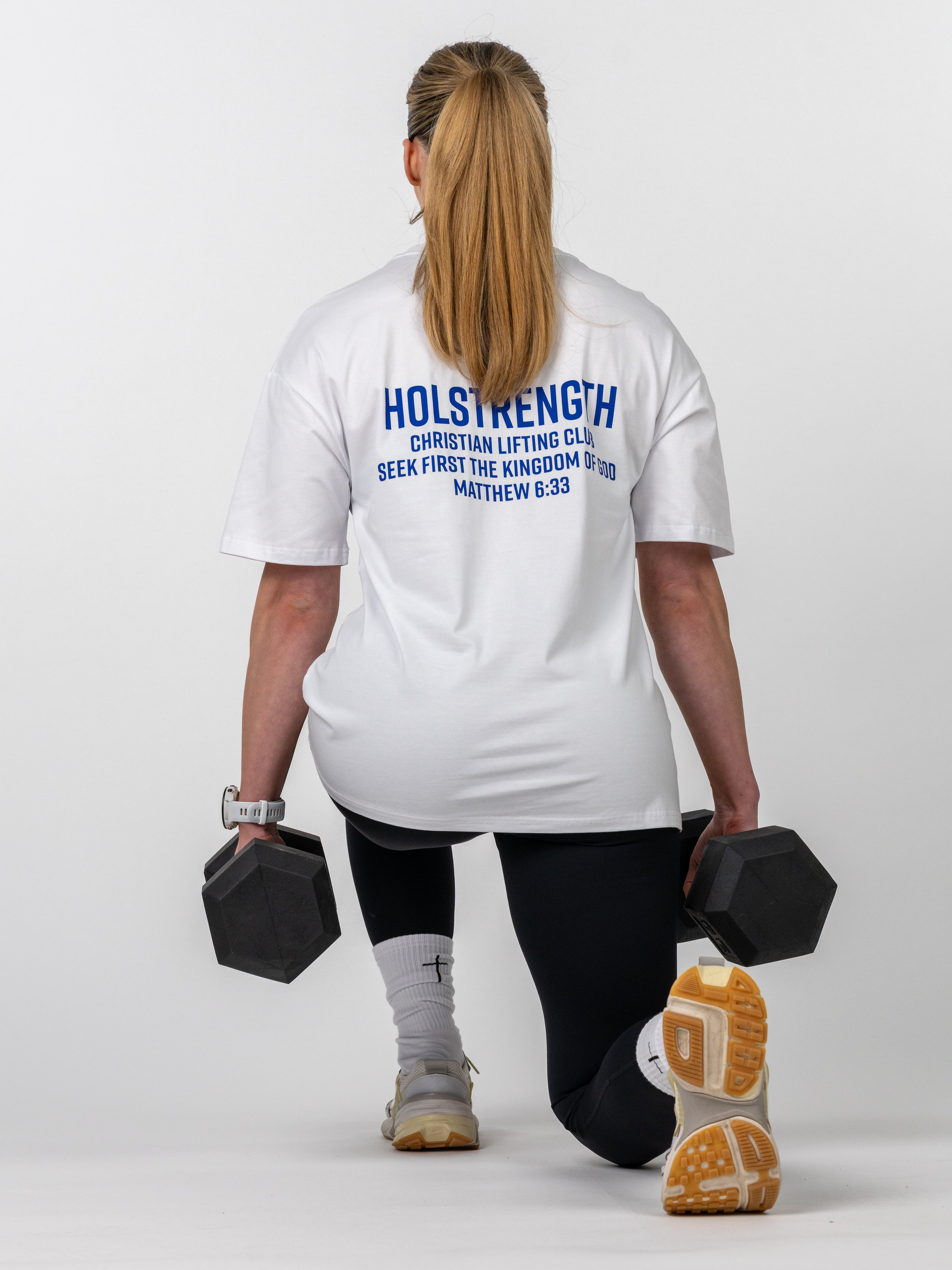 Woman performing a lunge exercise holding dumbbells wearing the Women's Lifting Club Oversized Tee with a faith-based fitness message on the back.