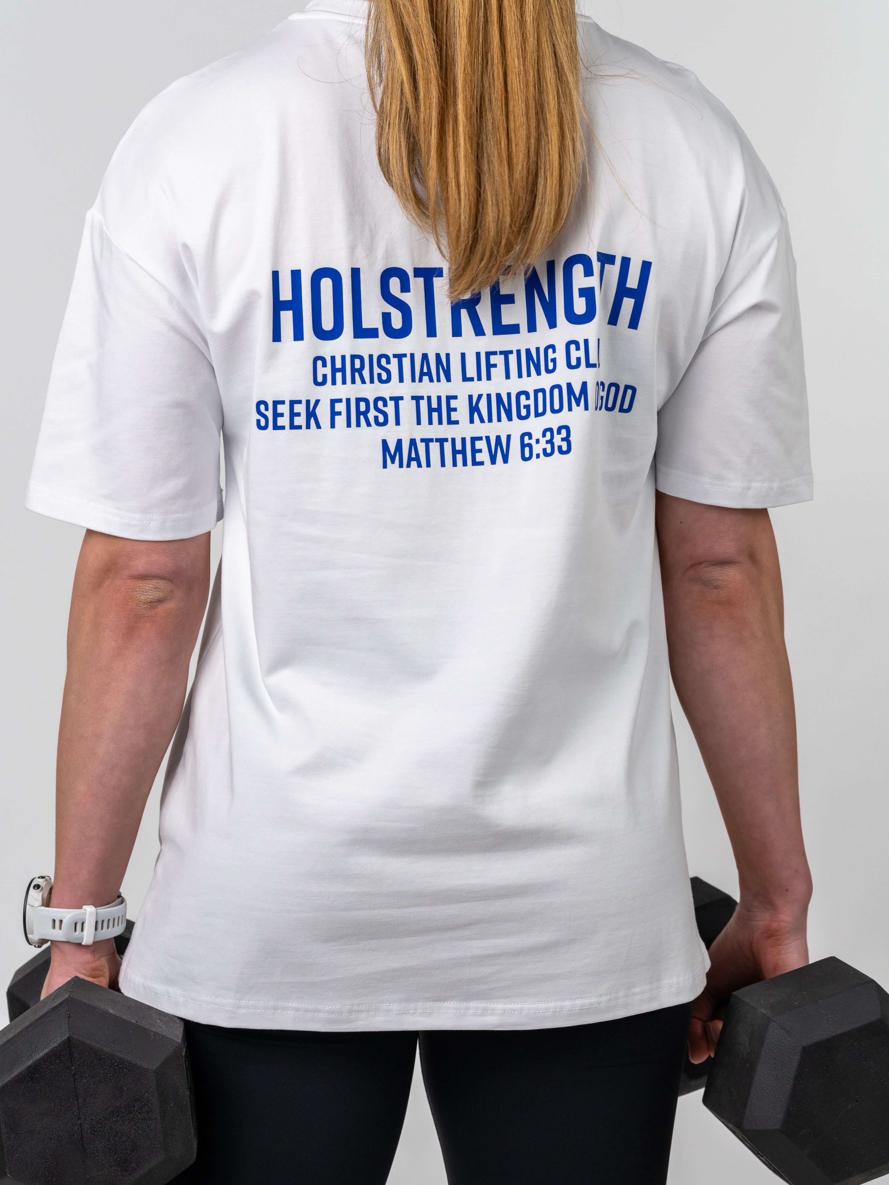 Woman wearing a Women's Lifting Club Oversized Tee with blue "Holstrength Christian Lifting Club Seek First the Kingdom of God Matthew 6:33" text on the back while holding black dumbbells.