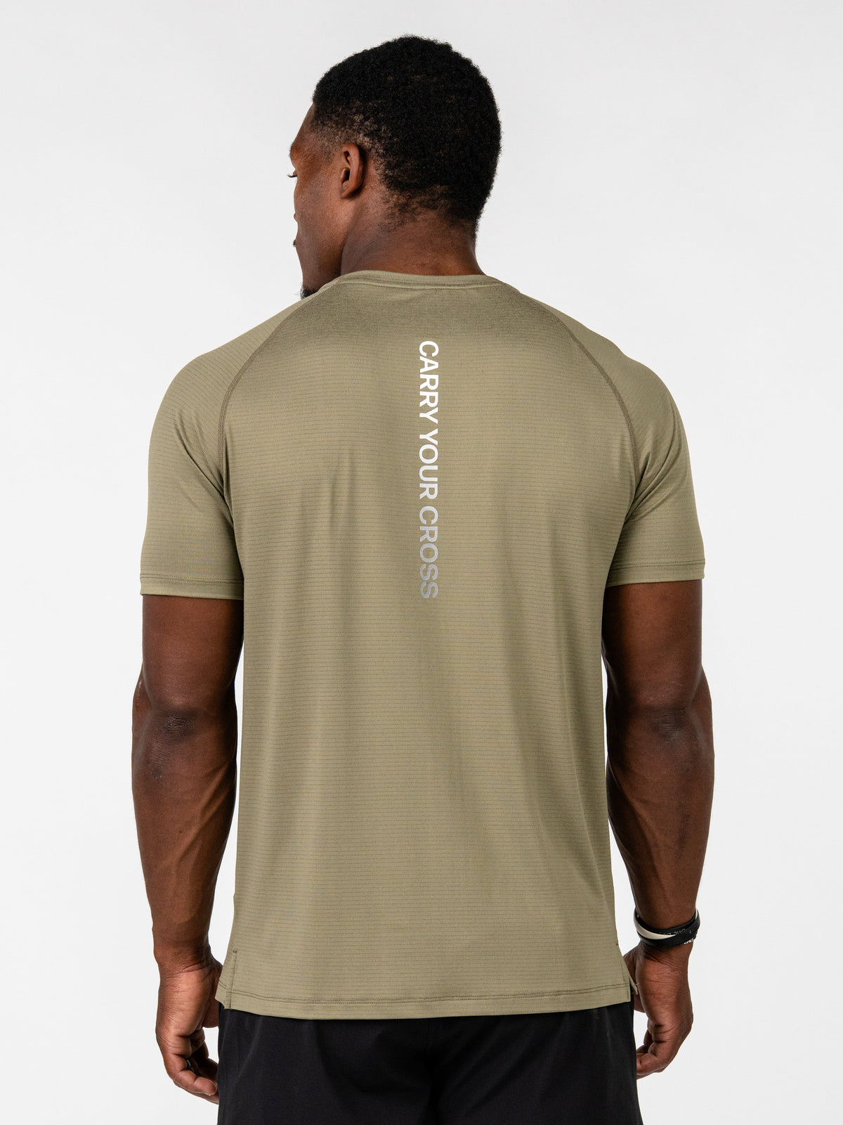 A man viewed from the back wearing a light khaki athletic t-shirt with 'CARRY YOUR CROSS' printed vertically in white on the back, paired with black athletic shorts, against a plain light gray background.