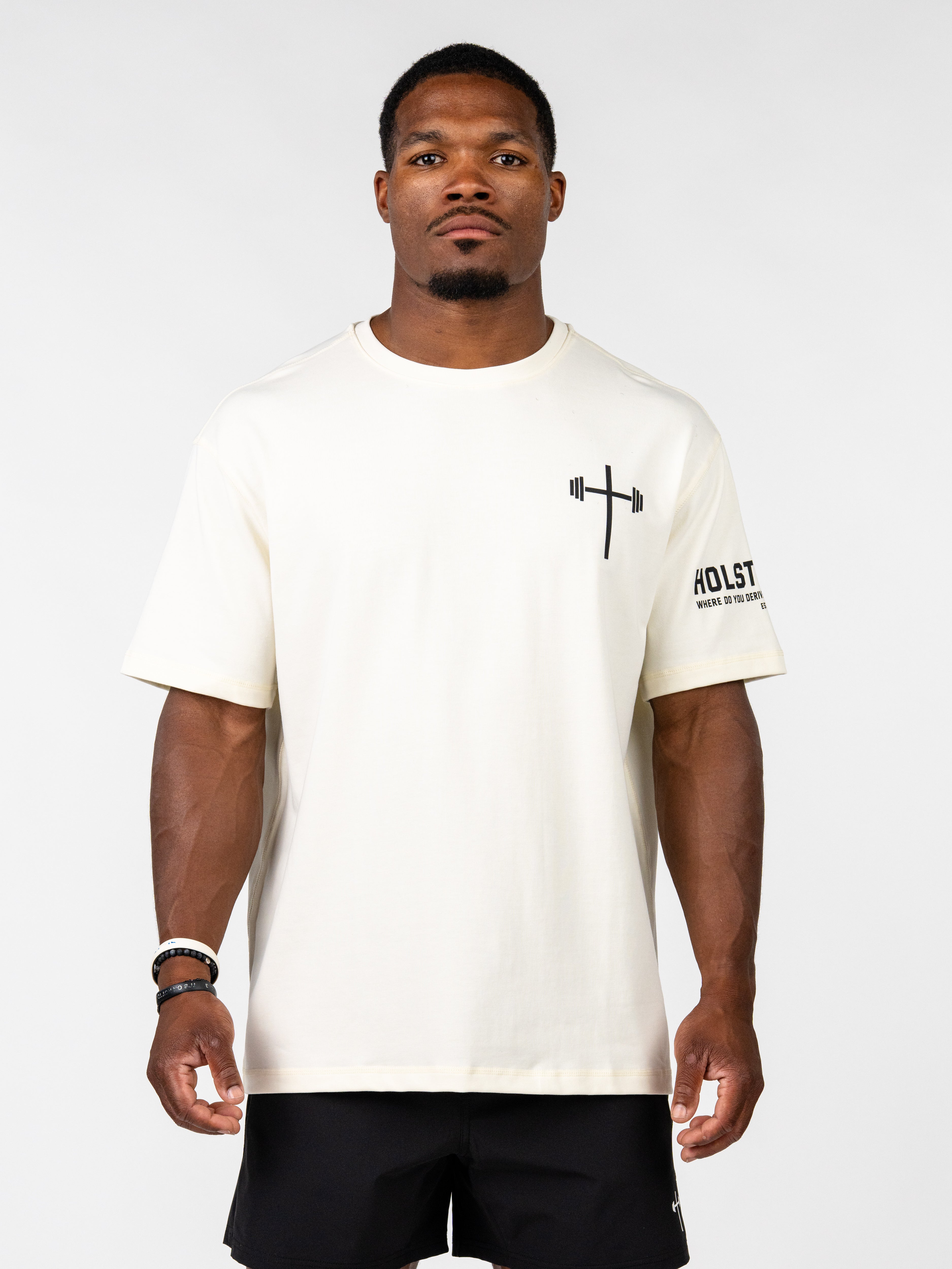 A man wearing a cream-colored short-sleeve t-shirt with a black barbell graphic on the left chest and black athletic shorts, standing against a light gray studio background.