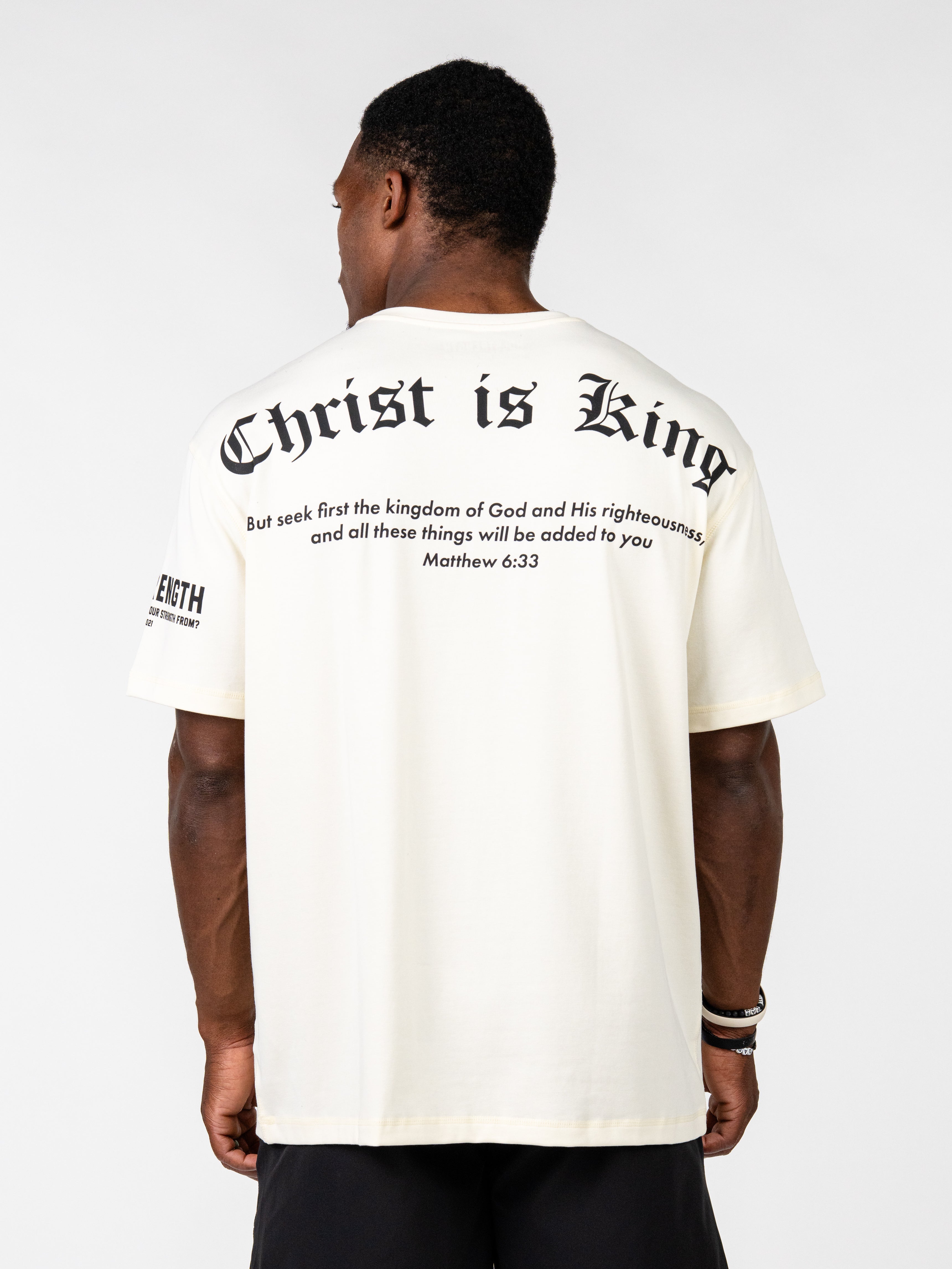 A man viewed from the back wearing an off-white t-shirt with black gothic text reading 'Christ is King' and scripture text on the back, paired with black shorts against a light gray studio background.