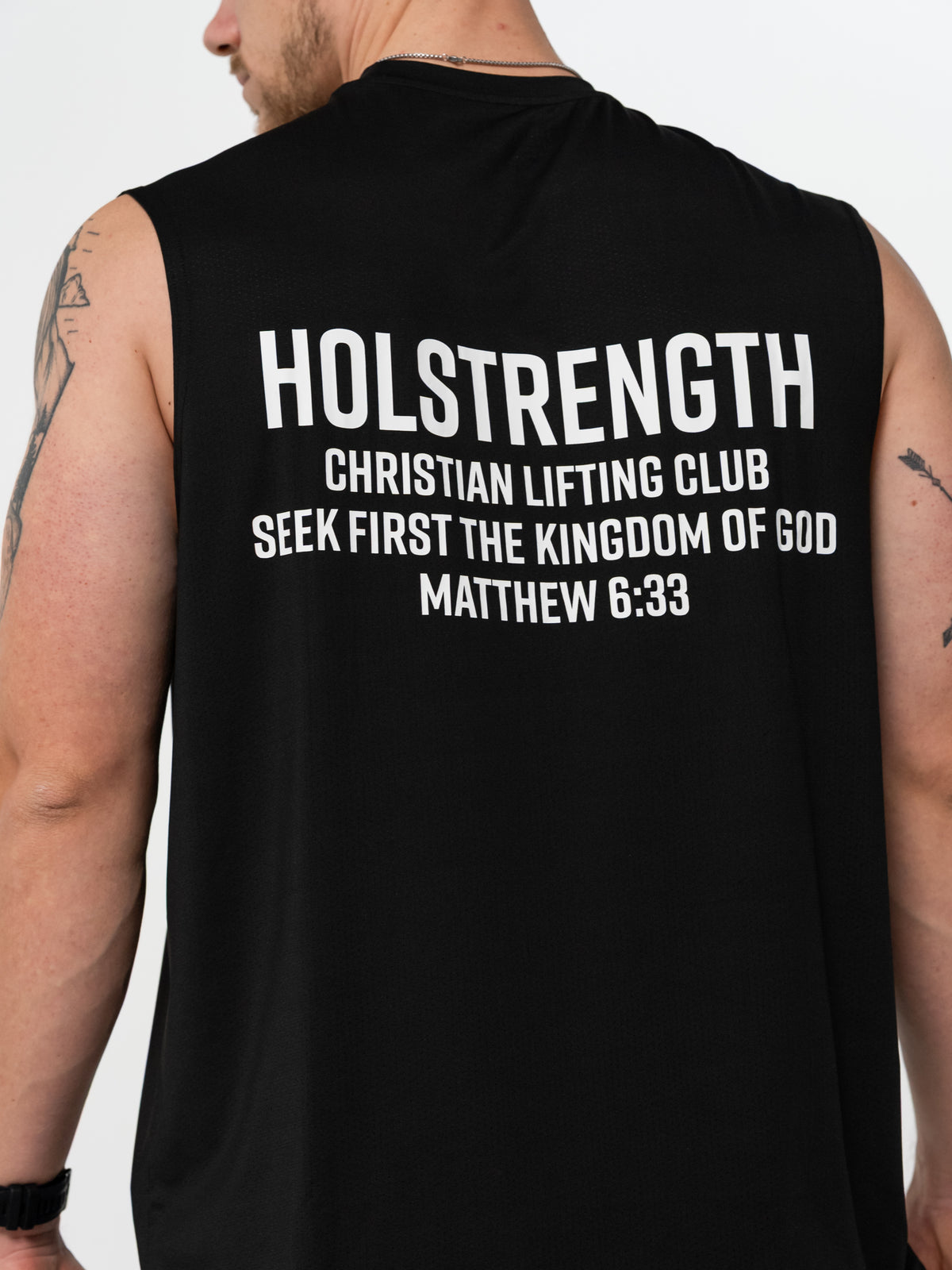 Back view of a person wearing a black sleeveless workout shirt with white text that reads Holstrength Christian Lifting Club Seek First the Kingdom of God Matthew 6:33, highlighting Christian gym clothing and faith-based fitness apparel.