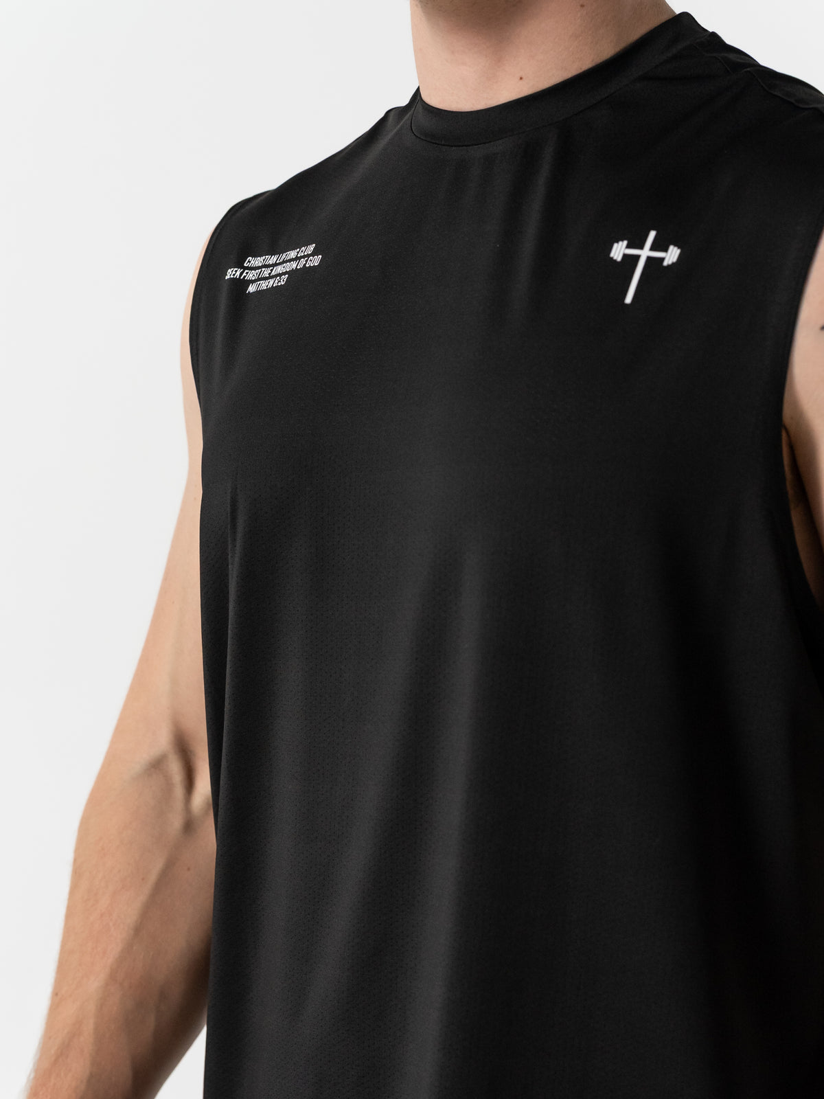 Close-up of a person wearing a black sleeveless workout shirt featuring a small white cross combined with a barbell icon on the upper chest, symbolizing Christian fitness apparel.