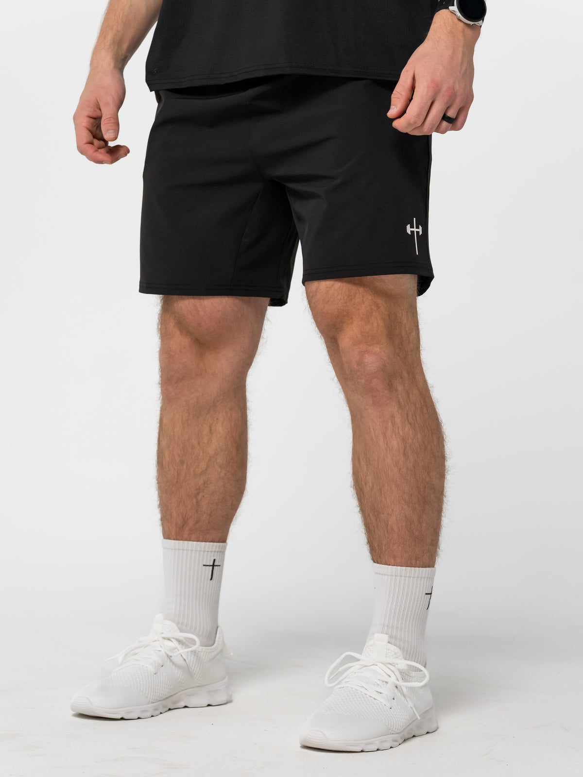 Man wearing black athletic shorts featuring a small white barbell cross logo on the left leg, paired with white socks and white sneakers, standing against a plain background.