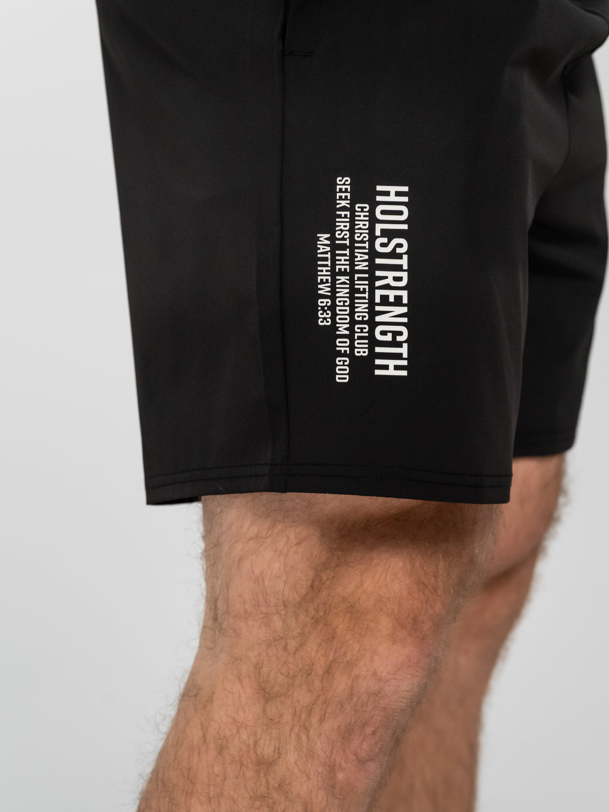 Close-up of a person's thigh wearing black shorts with white text that reads "HOLSTRENGTH Christian Lifting Club Seek First The Kingdom of God Matthew 6:33" on the side.