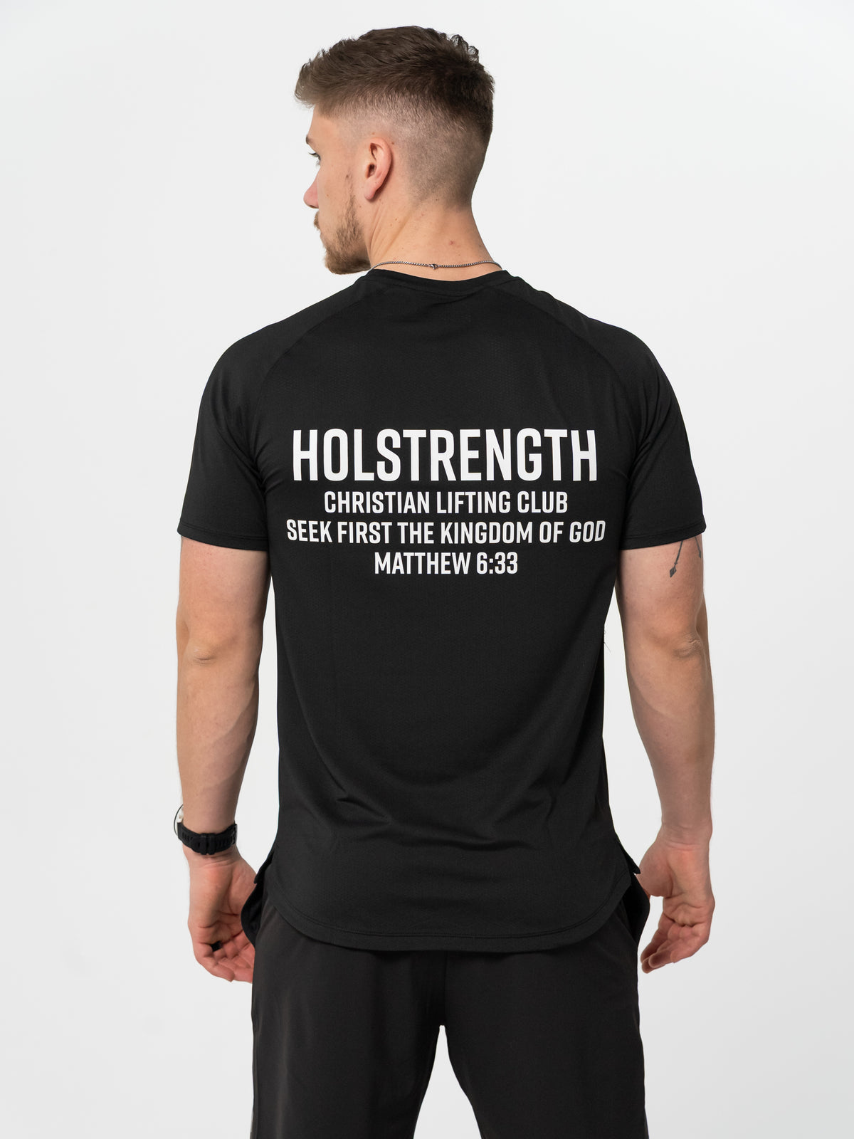 Man wearing black t-shirt and shorts, showing back of shirt with white text that reads "HOLSTRENGTH Christian Lifting Club Seek First The Kingdom of God Matthew 6:33," representing Christian fitness apparel.