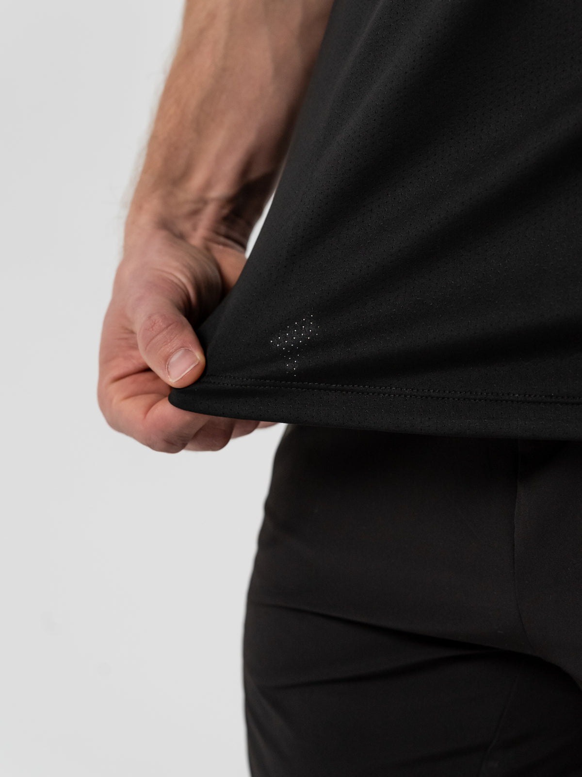 Close-up of a person wearing black athletic shorts and holding the edge of a black workout shirt, highlighting the fabric texture and subtle ventilation holes near the hem.