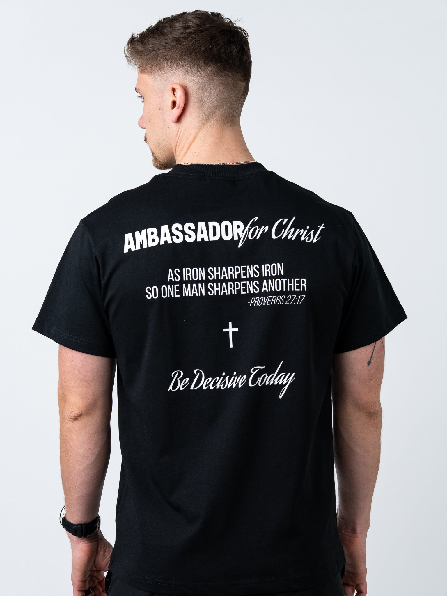 Person wearing a black t-shirt with text and a cross on the back, on a light gray background