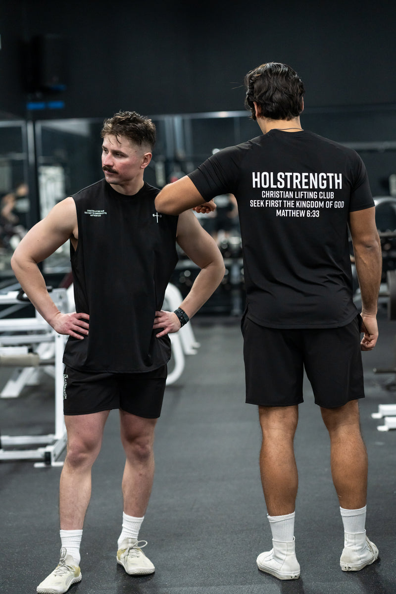 Two men wearing black sleeveless and short-sleeve Christian gym clothing stand in a gym with workout equipment. The shirt on the right displays "HOLSTRENGTH Christian Lifting Club Seek First the Kingdom of God Matthew 6:33" on the back.