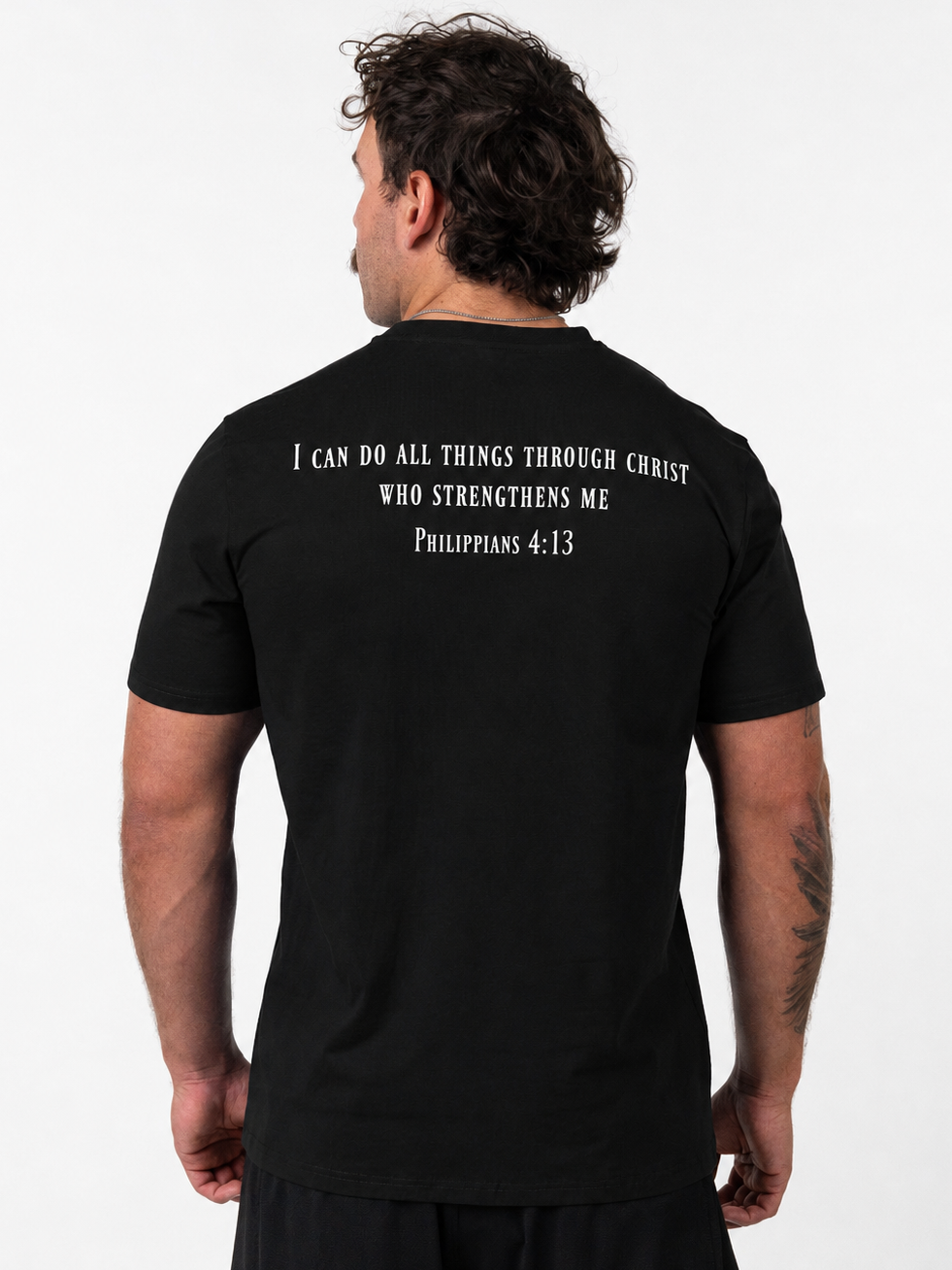 A man viewed from the back wearing a black t-shirt with white printed text on the upper back reading 'I CAN DO ALL THINGS THROUGH CHRIST WHO STRENGTHENS ME Philippians 4:13'.