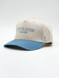 Beige baseball cap with a blue curved brim featuring embroidered text "GOD IS GOOD PSALM 23:6" on the front and a small blue cross stitched on the side.