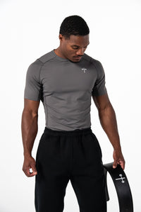 Athletic man wearing a fitted gray Christian fitness apparel short-sleeve shirt with a small cross and barbell logo on the chest, holding a black lifting belt with the same logo.