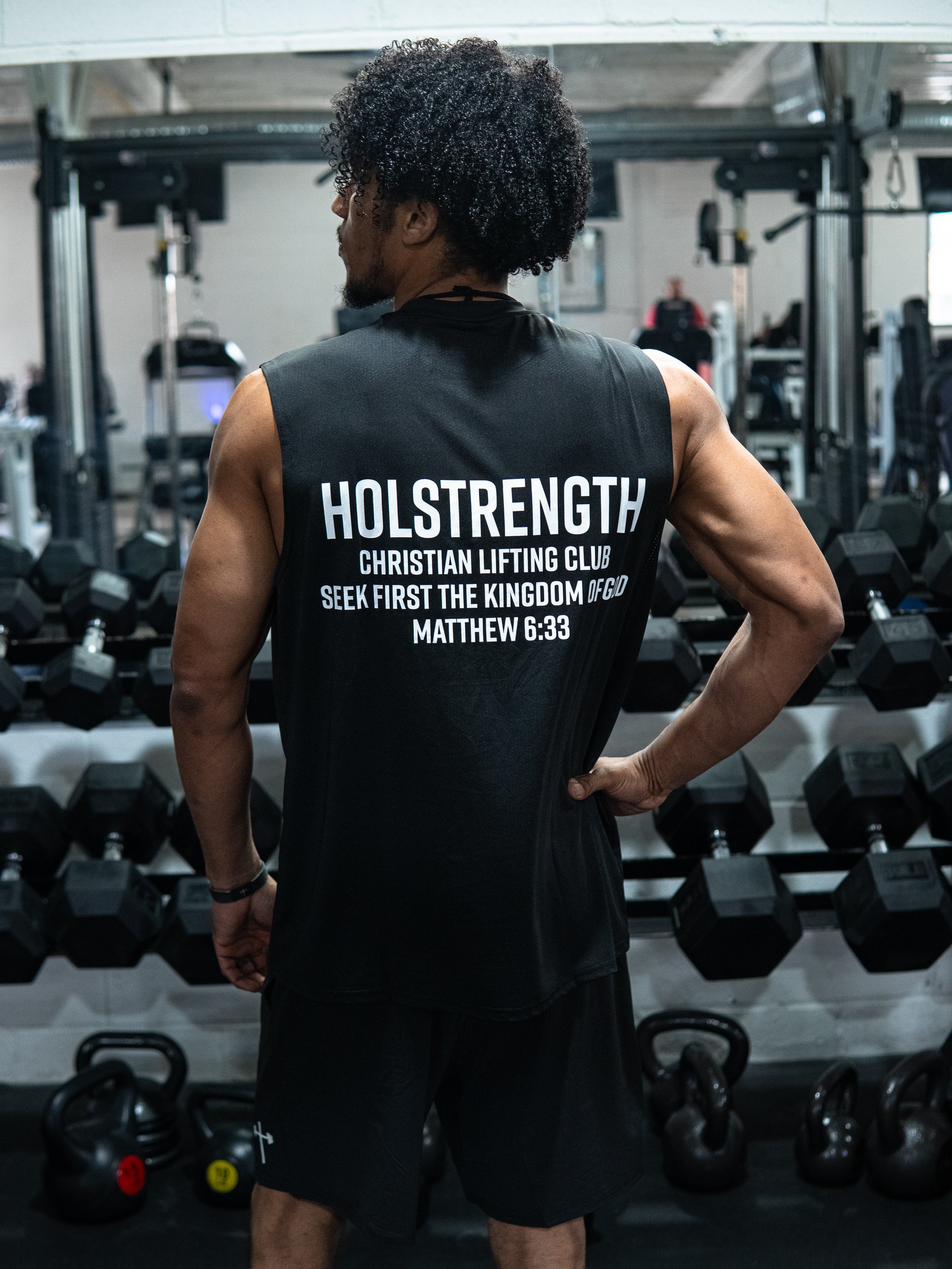 Man wearing Christian Lifting Club Premium Oversized Tank with faith-based motivational text on back, standing in gym surrounded by weights and kettlebells.
