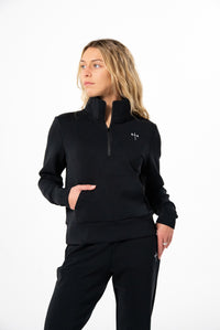 Woman wearing a black quarter-zip pullover and matching black pants with a small white barbell cross logo on the chest and thigh, standing against a white background.