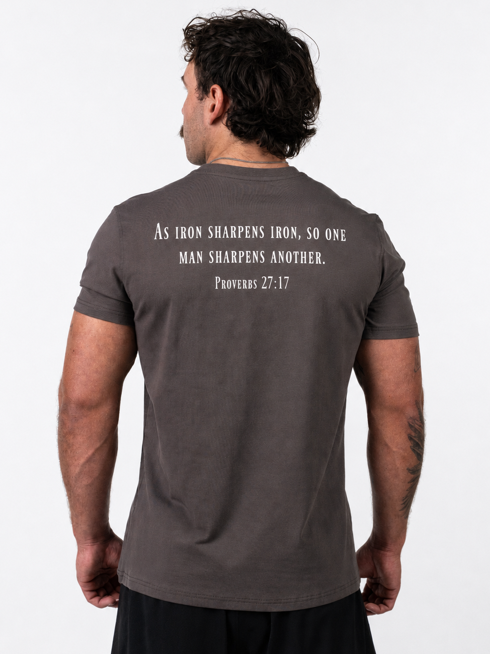 A man viewed from behind wearing a dark brown t-shirt with a white printed biblical quote on the back, paired with black shorts, against a plain white background.