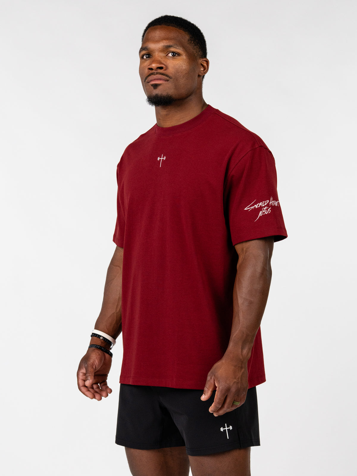 Sacred Heart Premium Oversized Tee in deep red worn by a male model with a small cross chest emblem and sleeve script with black athletic shorts