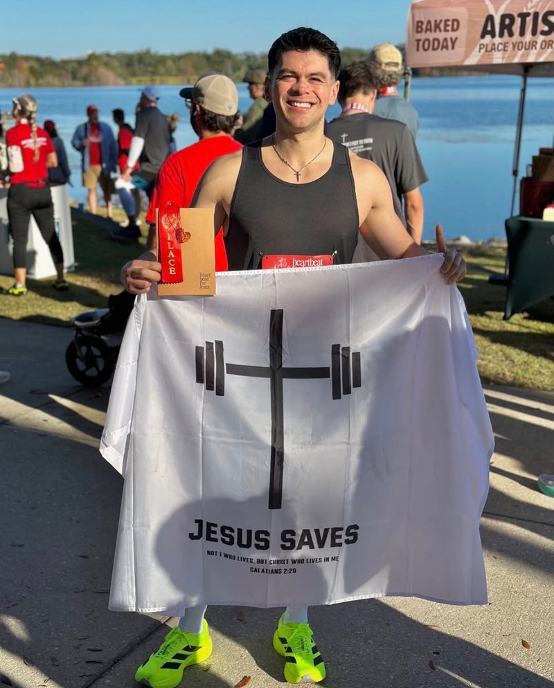 Man wearing a black tank top and bright yellow shoes holding a white flag with a cross-shaped barbell and the words "JESUS SAVES" along with a Bible verse, standing outdoors near a lake with other people in the background.