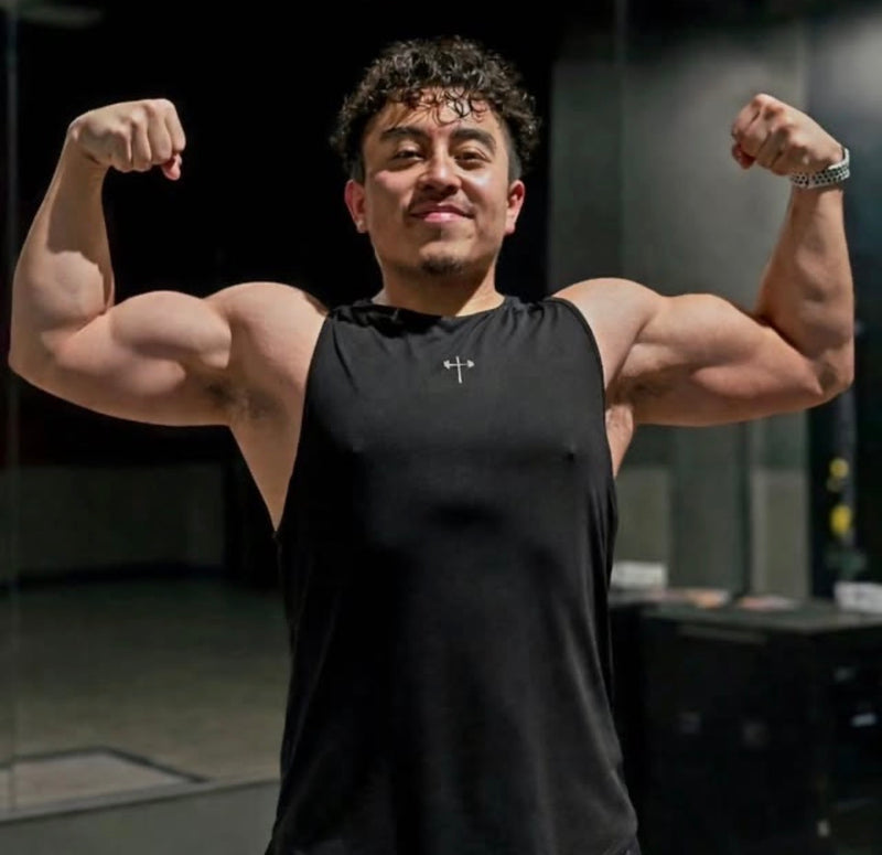 Man wearing sleeveless black workout shirt with a small cross design flexing his biceps in a gym setting with a dark background.