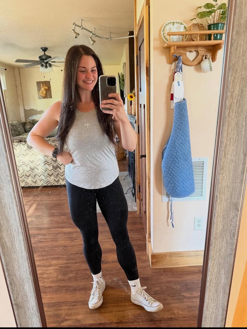Woman taking a mirror selfie wearing a light gray sleeveless top with a small cross design, black leggings, and white sneakers in a cozy home setting.