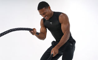 Athletic man wearing a black sleeveless shirt and pants, engaged in intense battle rope workout with visible muscle definition against a white background.