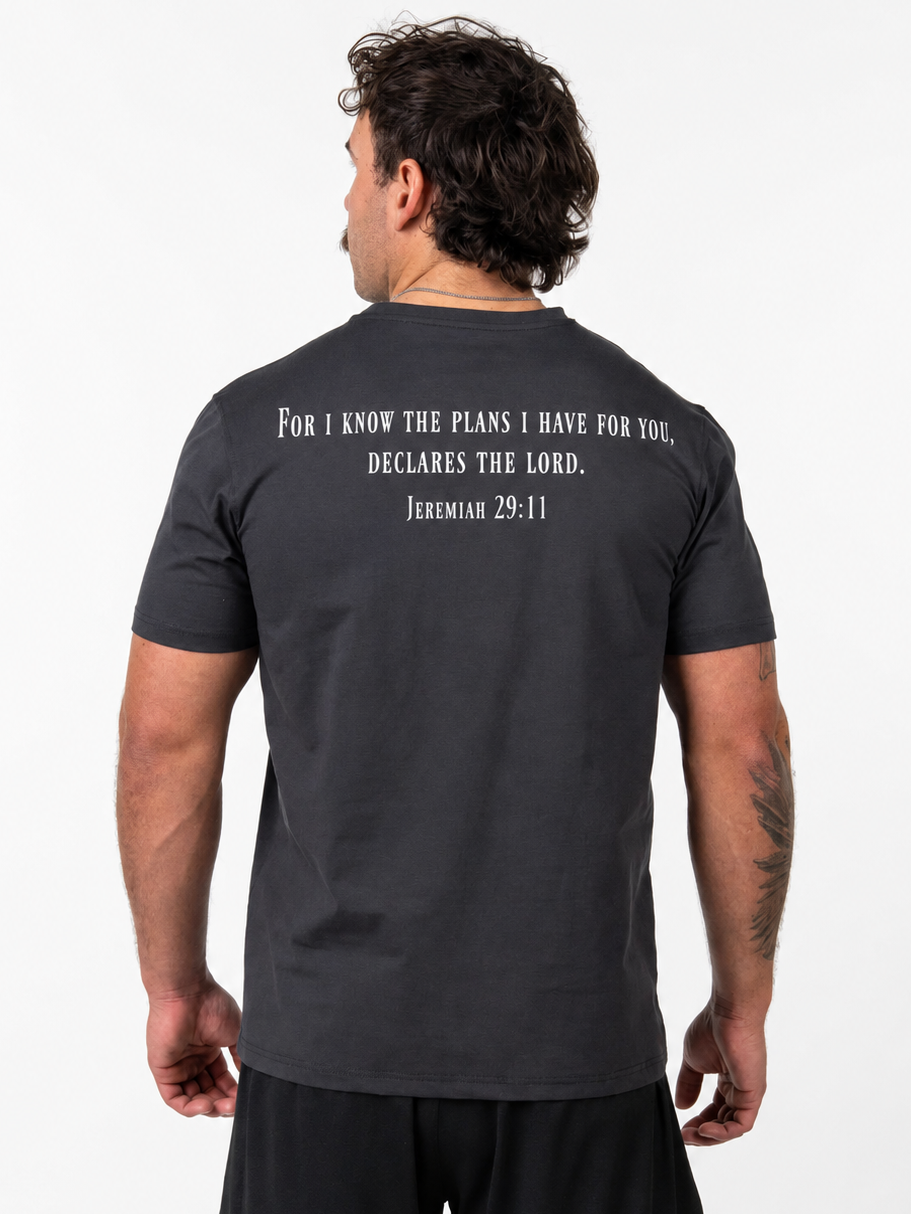 A man viewed from the back wearing a dark charcoal gray t-shirt with a white printed Bible verse from Jeremiah 29:11 on the upper back, paired with black athletic shorts, against a plain white background.