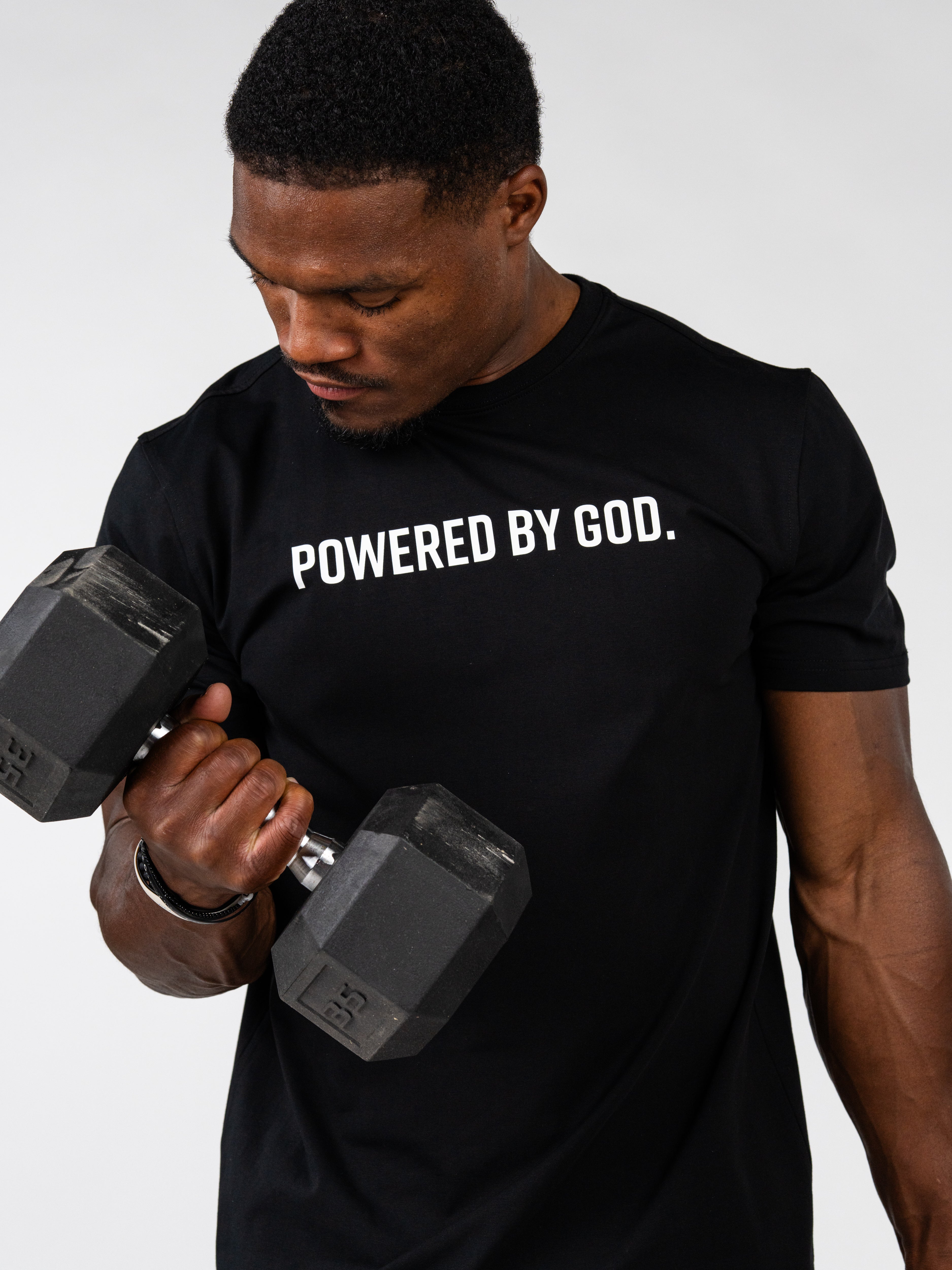 A man lifting a hex dumbbell while wearing a black t-shirt with the white printed text 'POWERED BY GOD.' on the chest, set against a plain light gray background.