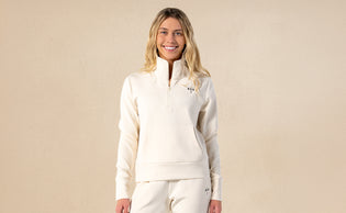 Smiling woman wearing a cream-colored quarter-zip pullover and matching pants standing against a plain beige background.