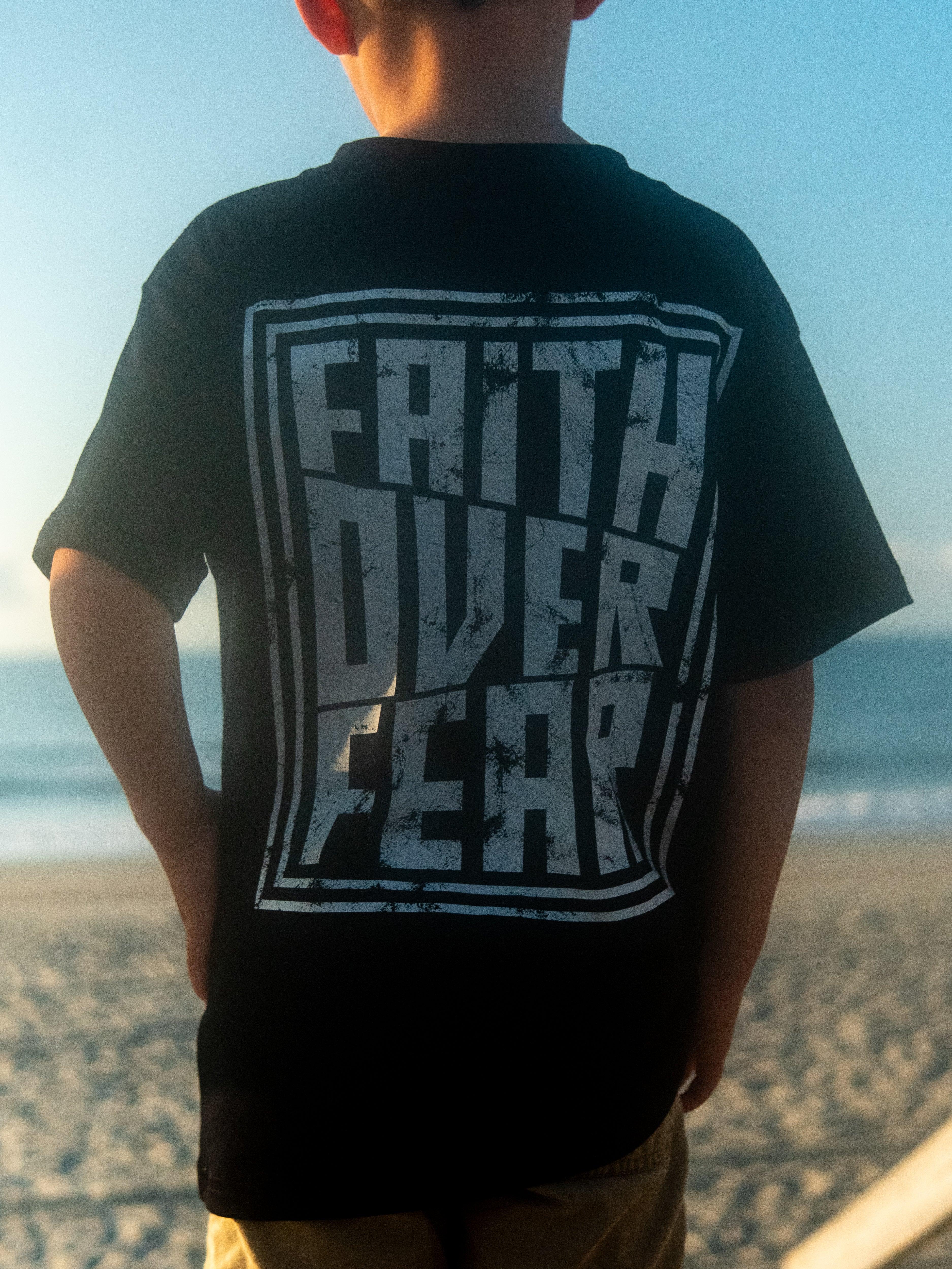 Child wearing Faith Over Fear Youth Tee with bold "Faith Over Fear" graphic on the back, standing on a beach with ocean in the background, Christian gym clothing style.