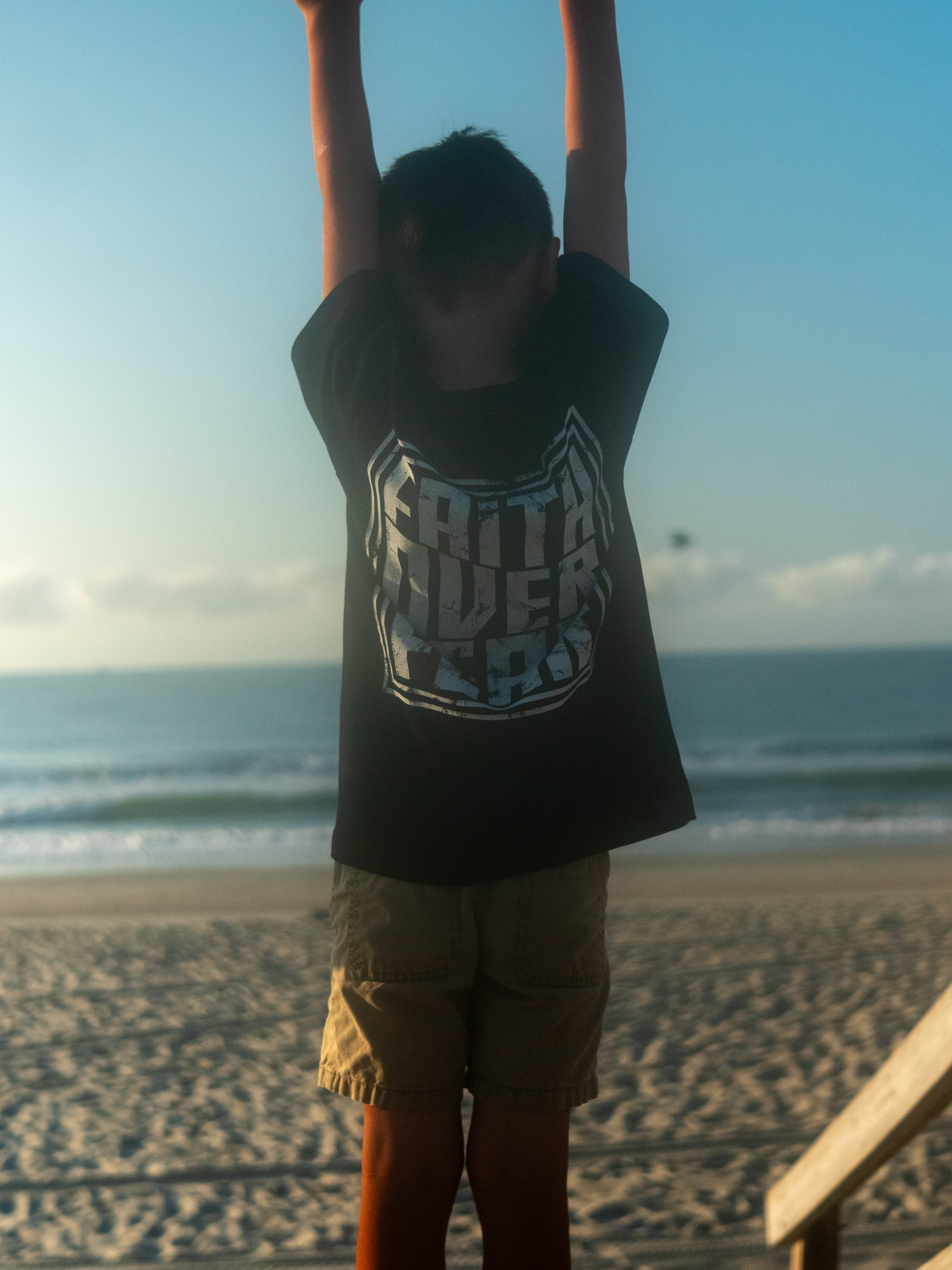 Child wearing a Faith Over Fear Youth Tee with an oversized graphic on the back, standing with arms raised at a beach during sunrise or sunset.
