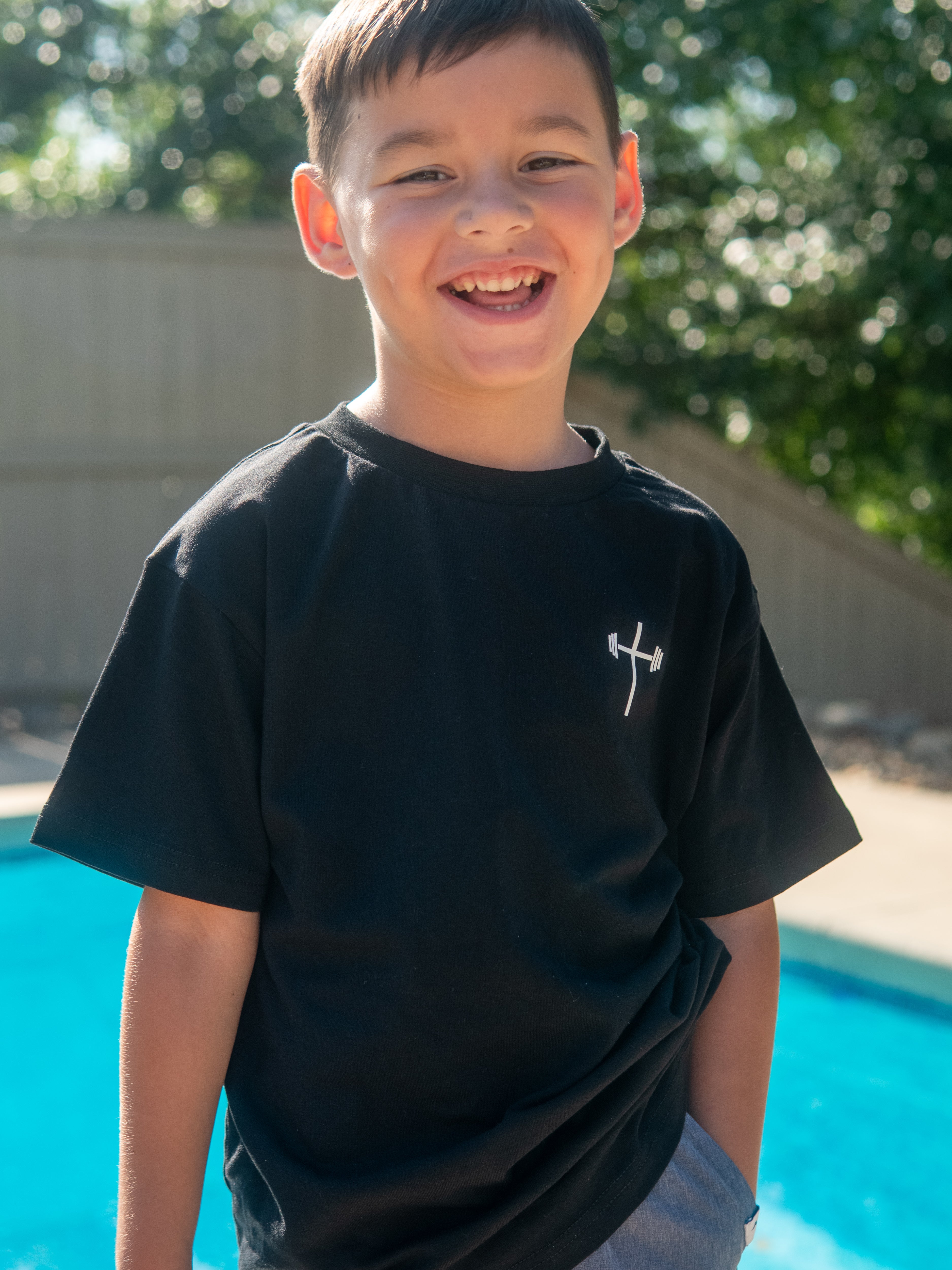 Young boy smiling outdoors wearing a black Jesus is King Youth Tee featuring a small white barbell cross logo on the front, styled as Christian gym clothing.