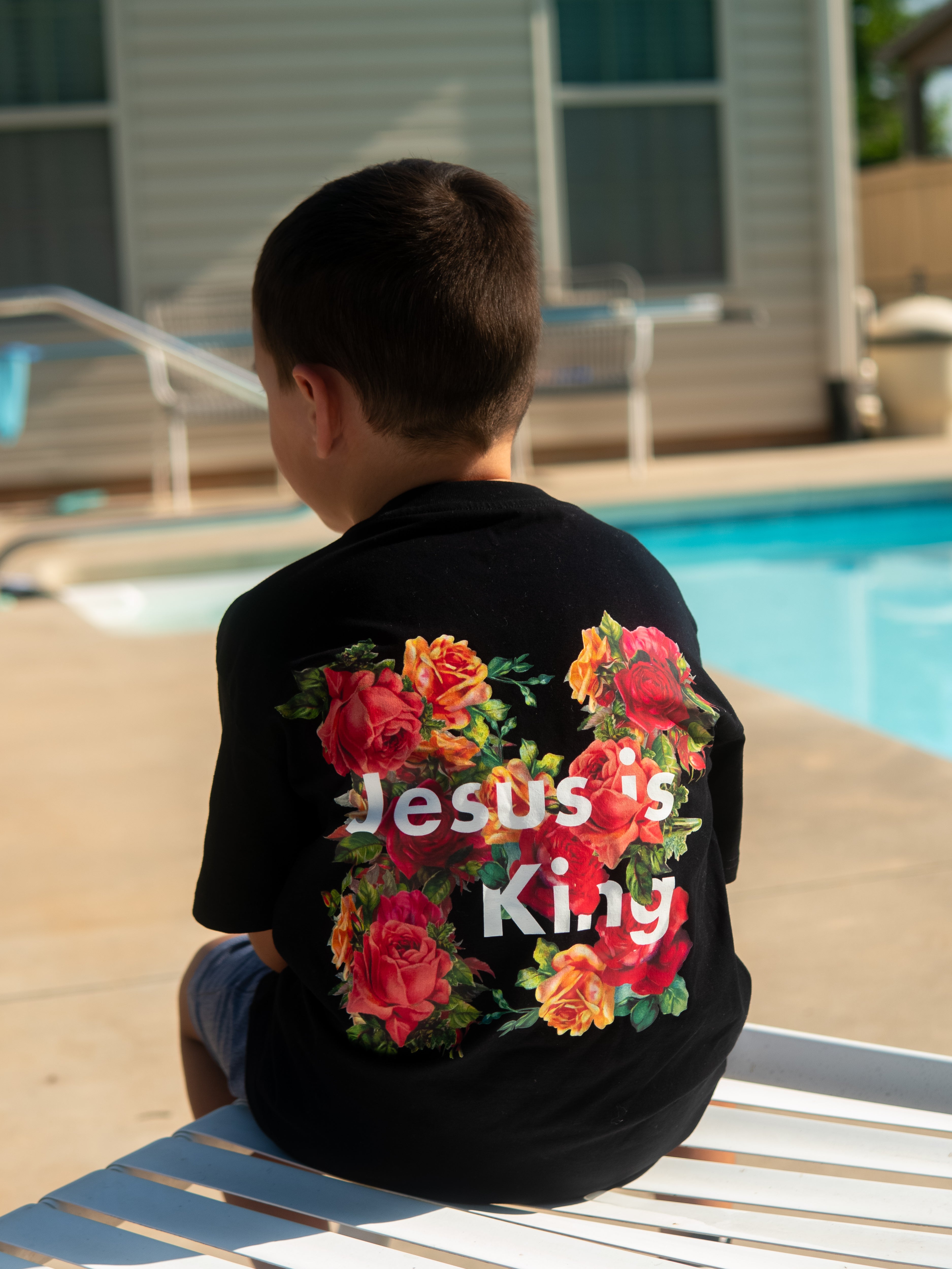 Youth sitting by pool wearing Jesus is King Youth Tee featuring colorful floral graphic and bold white text on black fabric, representing Christian fitness apparel.