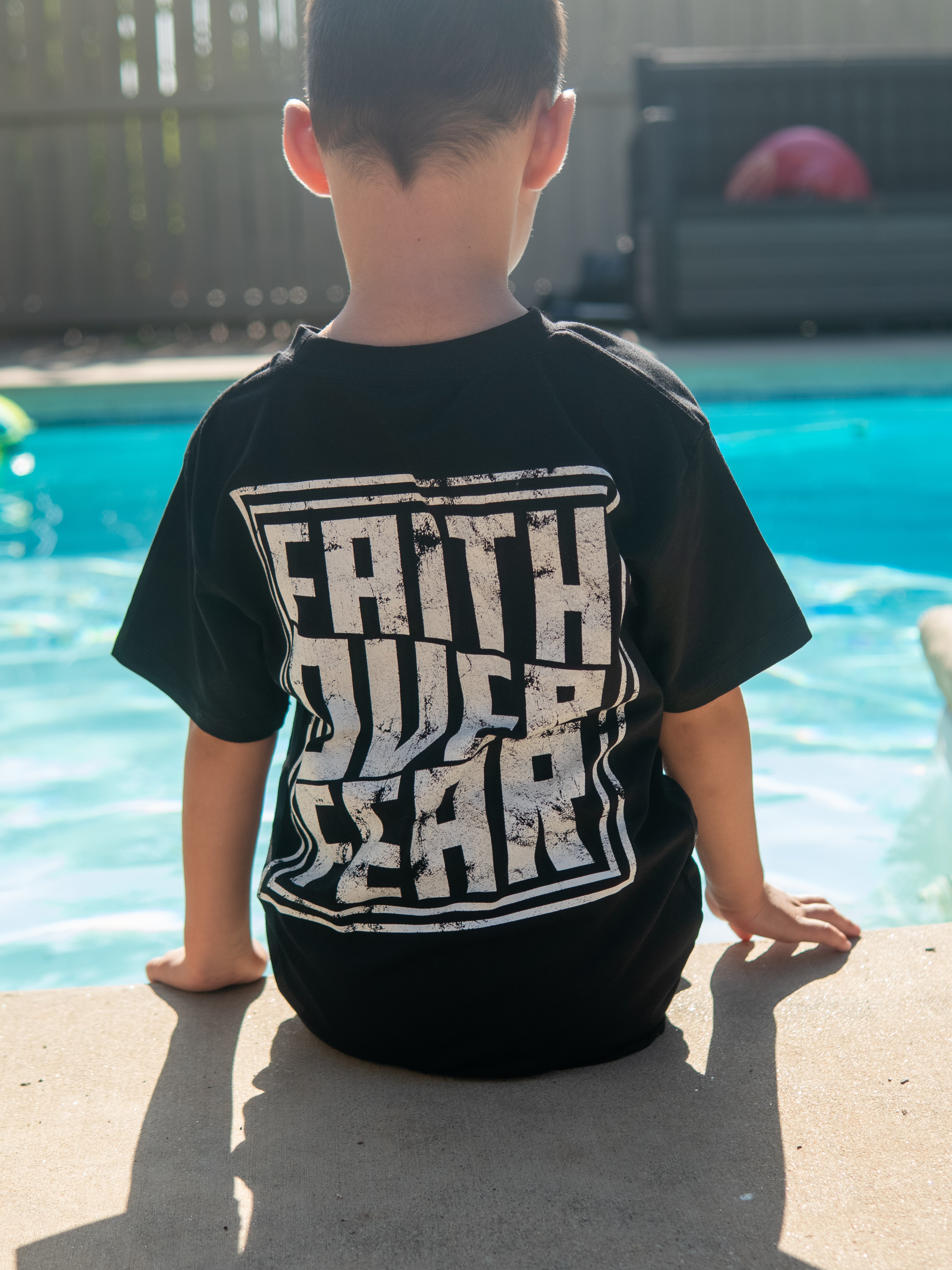 Young boy sitting by a pool wearing a black Faith Over Fear Youth Tee with a large white graphic text on the back promoting faith based apparel.