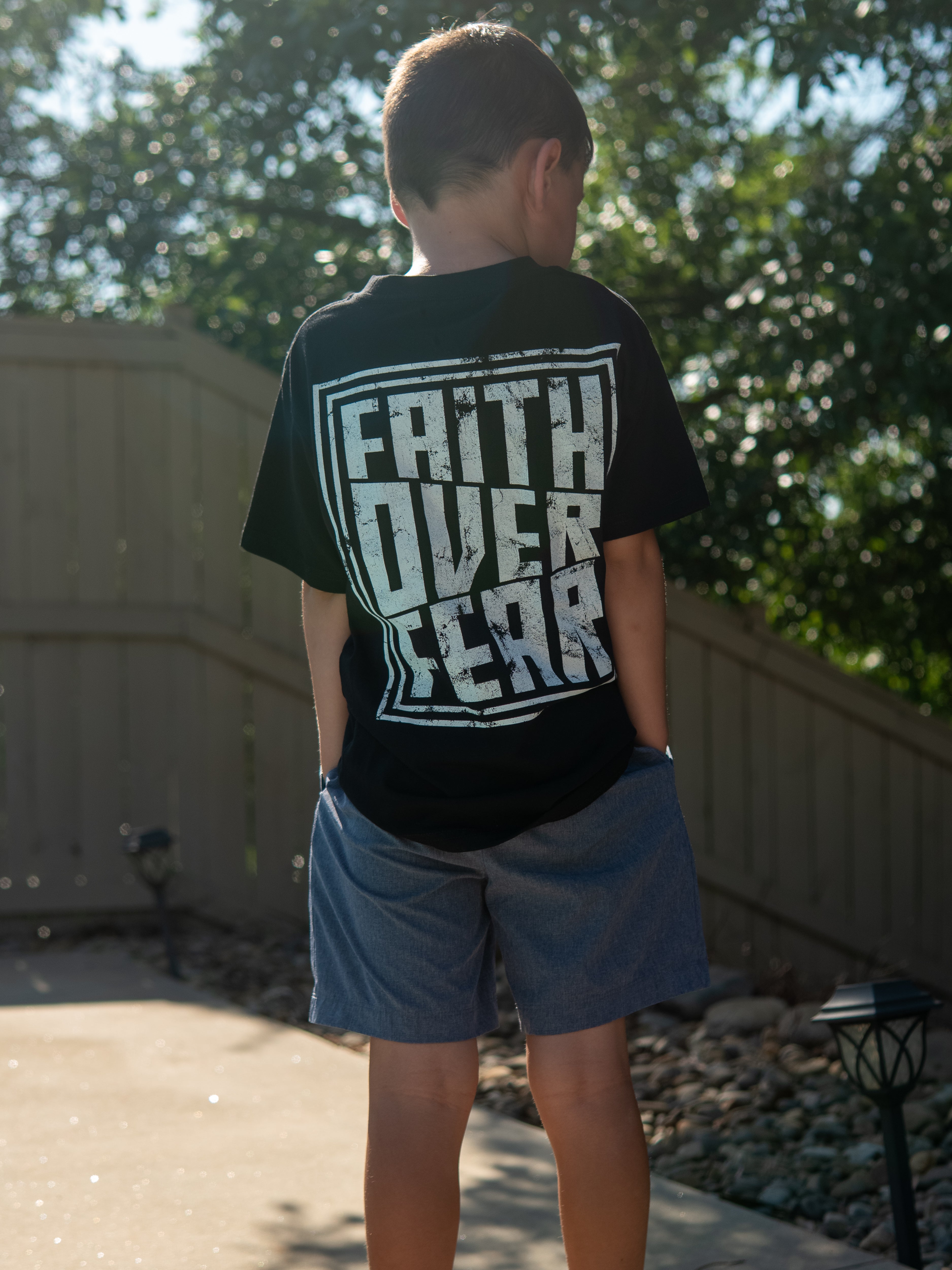Young boy standing outdoors wearing a black Faith Over Fear Youth Tee with bold white graphic text on the back, paired with gray shorts in a casual setting.