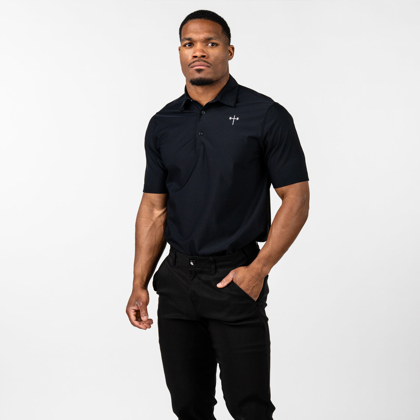 Man wearing a fitted black short-sleeve polo shirt with a small white cross logo on the left chest, paired with black pants, standing against a plain white background.