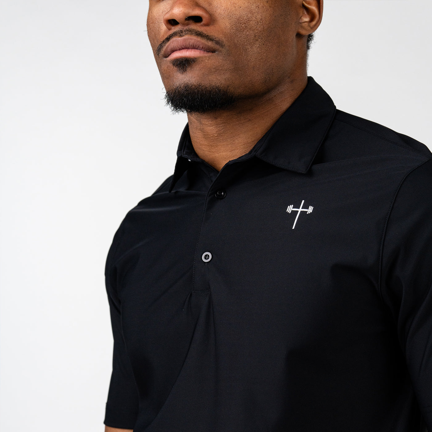 Close-up of a man wearing a black athletic polo shirt featuring a small white barbell cross logo on the left chest against a plain white background.