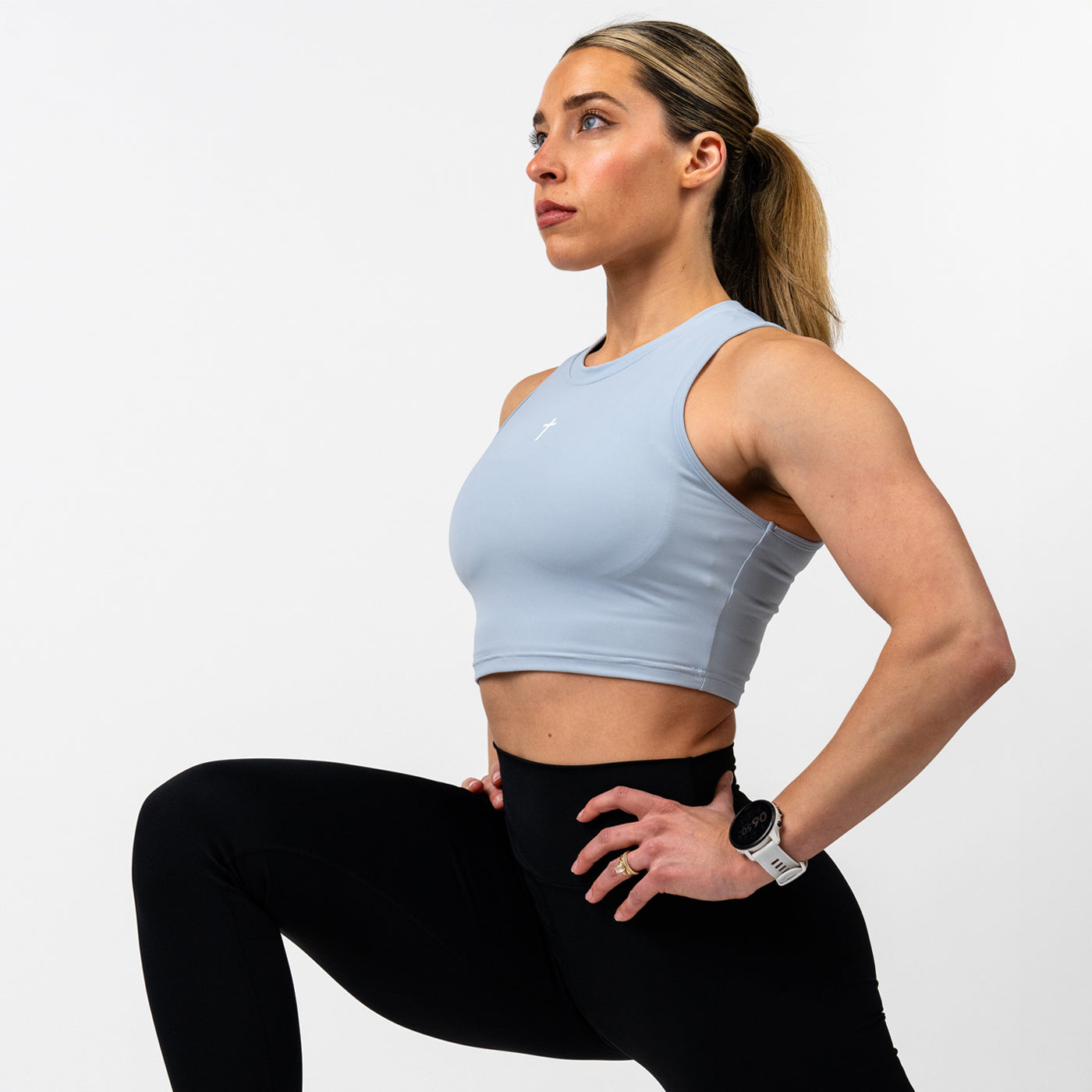 Woman in athletic wear performing a lunge exercise, wearing a light blue sleeveless crop top with a subtle cross symbol and black high-waisted leggings.