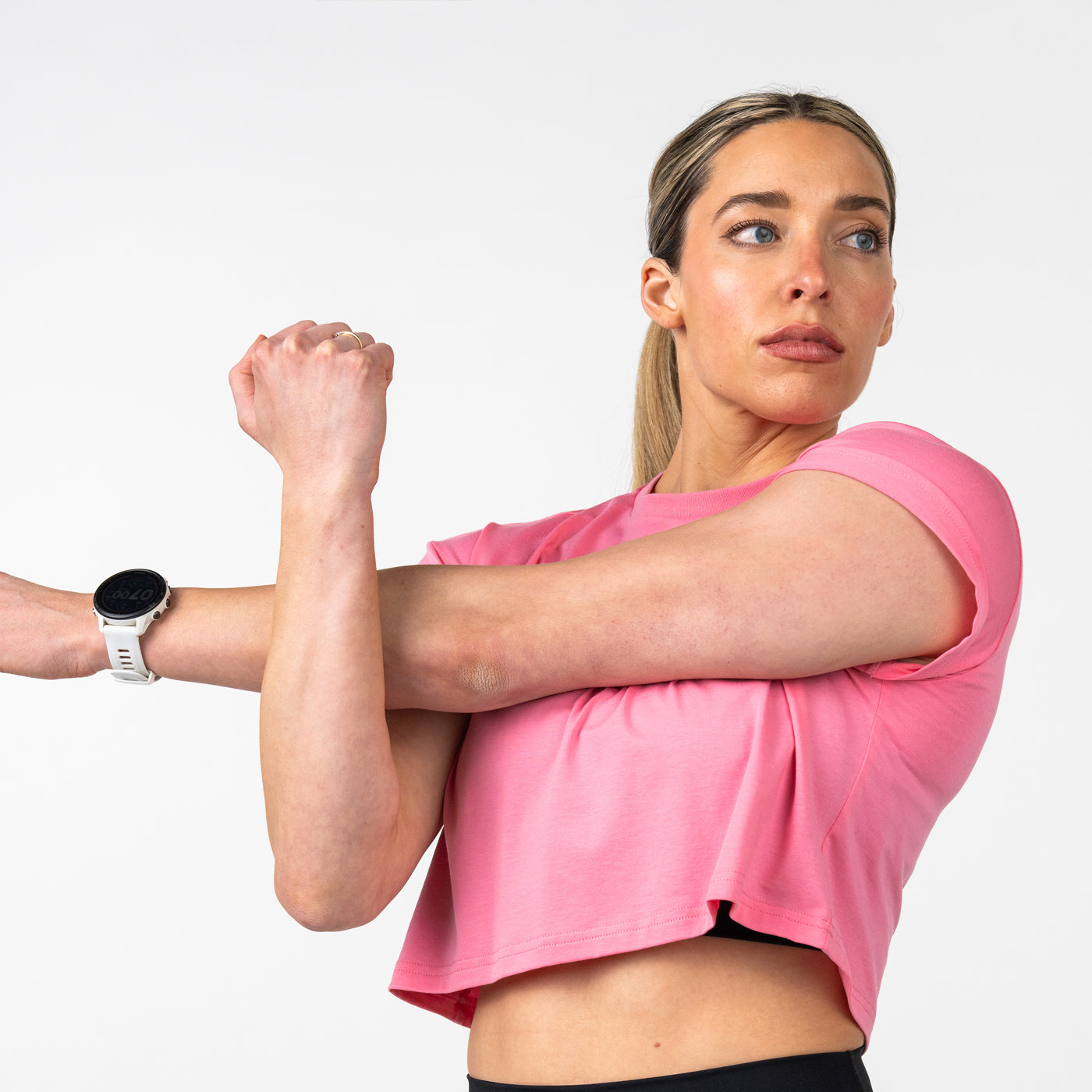 Athletic woman with blonde hair wearing a pink short-sleeve crop top stretching her arm, wearing a white sports watch against a plain white background.