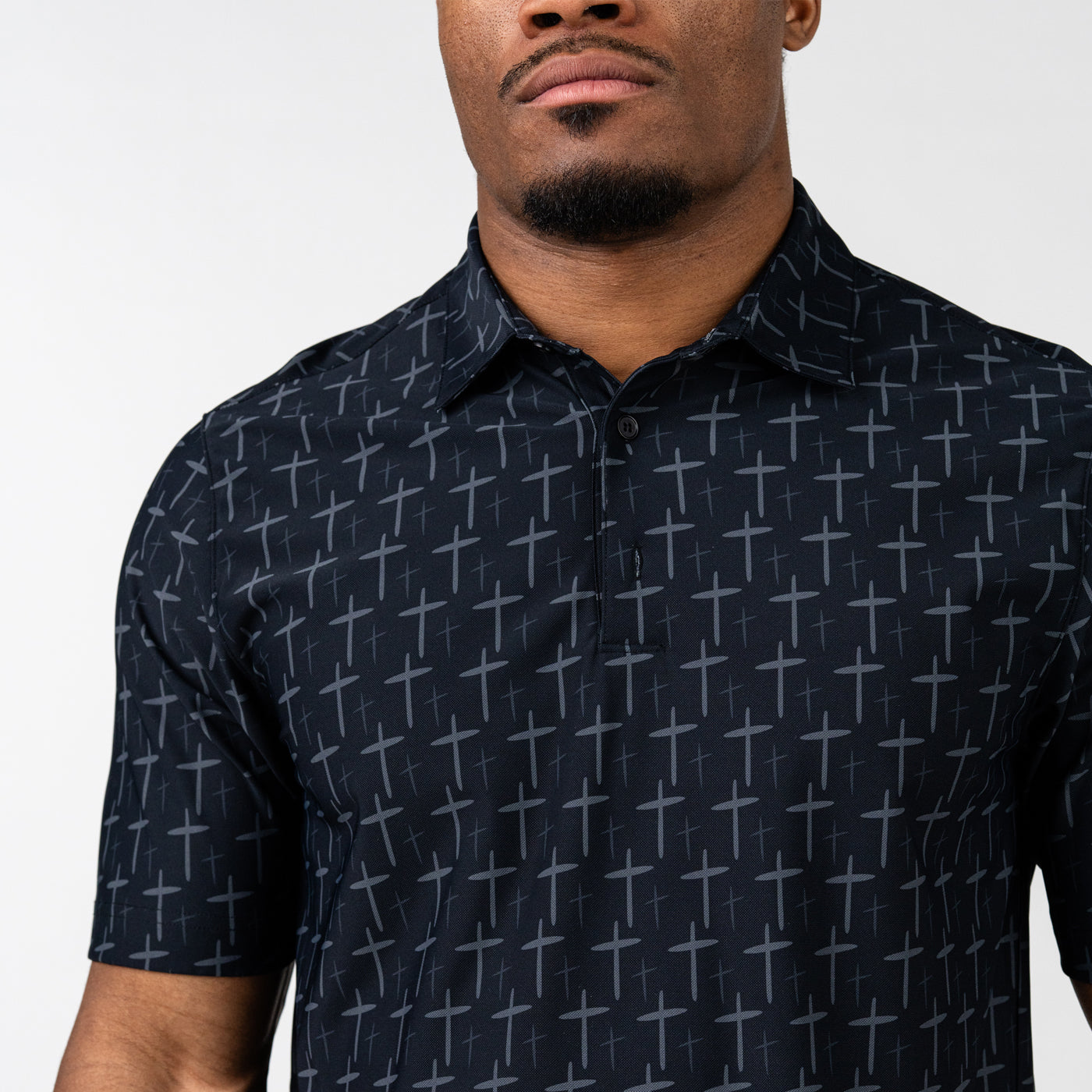 Close-up of a man wearing a black polo shirt with a subtle all-over pattern of gray crosses, featuring a collar and a button placket.