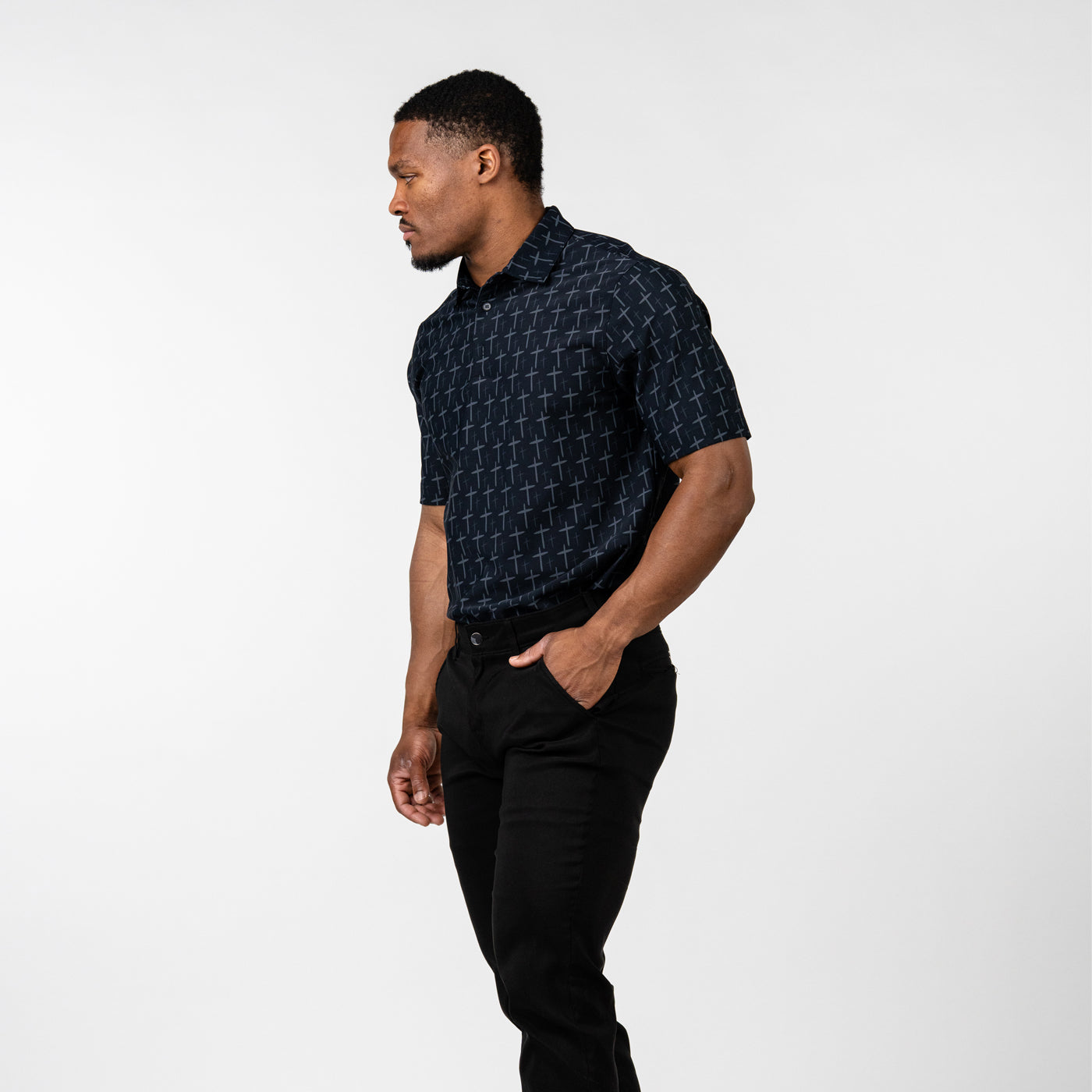 Athletic man standing in profile wearing a black short-sleeve polo shirt with a subtle cross pattern and black pants against a plain white background.