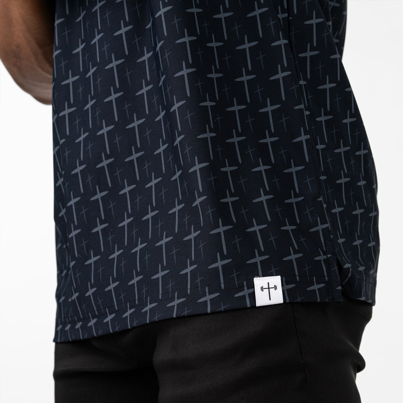 Close-up of a black athletic shirt featuring a repeating barbell cross pattern and a small logo patch near the hem, worn with black pants.