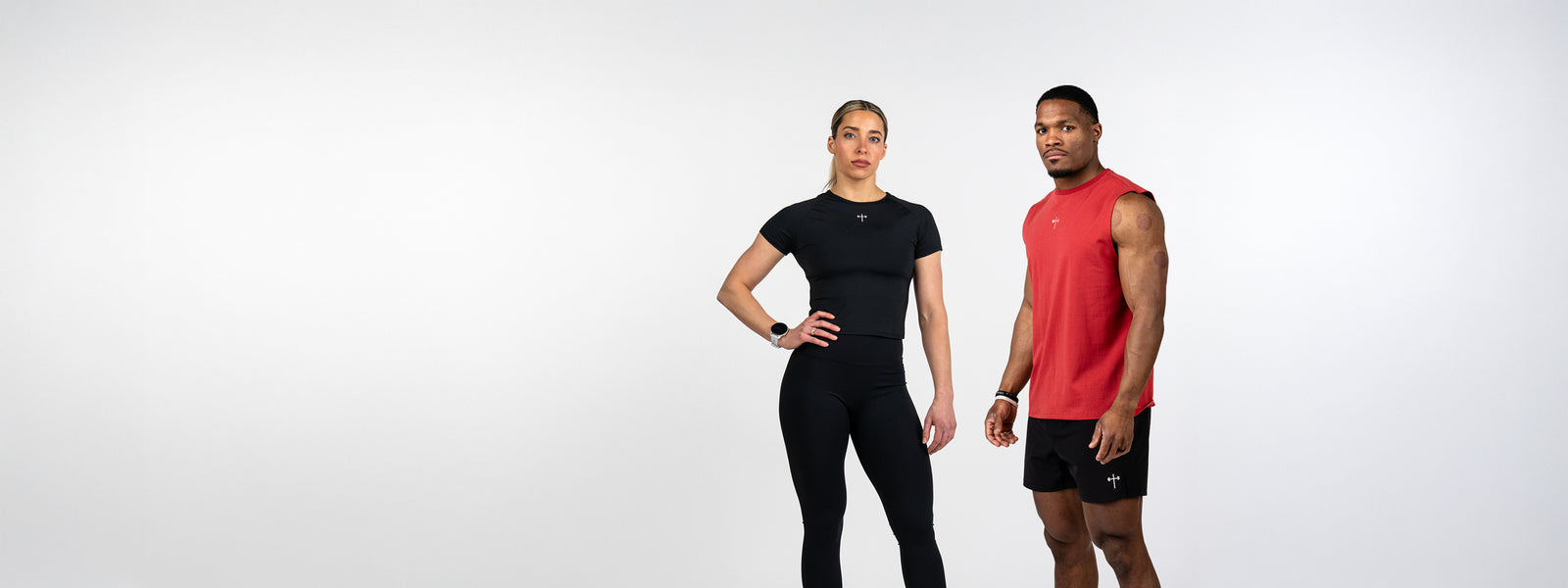 Two fitness models wearing athletic clothing stand against a plain white background, one in a black short-sleeve top and leggings, the other in a red sleeveless shirt and black shorts.