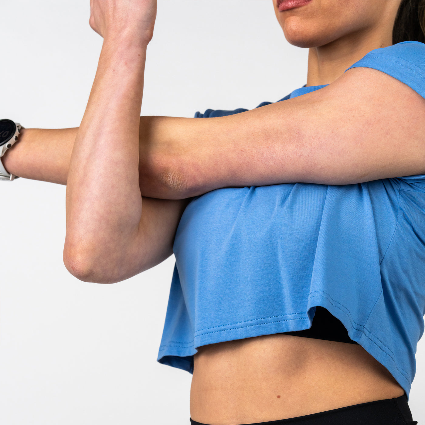 Close-up of a person in a light blue cropped workout top stretching their arm across their chest, wearing a black fitness watch on the wrist, against a plain white background.