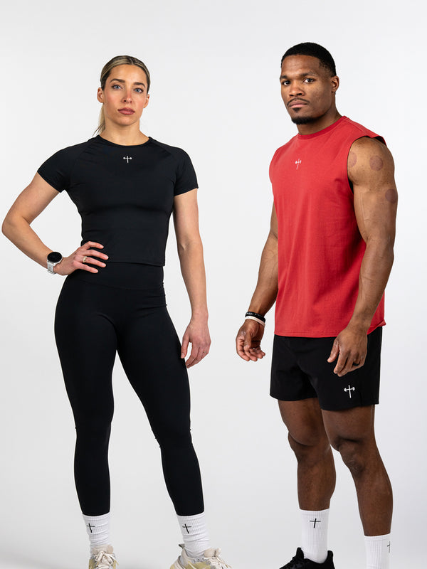 A woman and man standing side by side wearing Christian fitness apparel; the woman is dressed in black leggings and a black short-sleeve crop top, and the man is wearing black shorts and a red sleeveless shirt, both outfits featuring small cross logos.