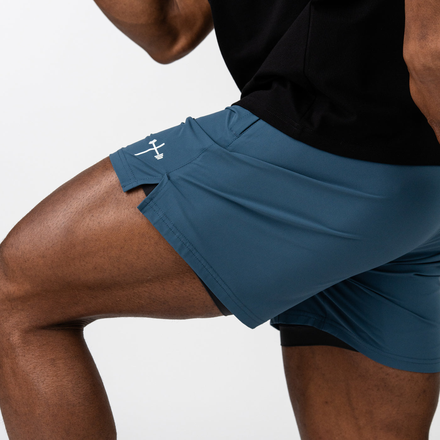 Close-up of a person wearing blue athletic shorts with a white barbell cross logo near the hem, paired with a black top, highlighting muscular legs in a fitness pose.
