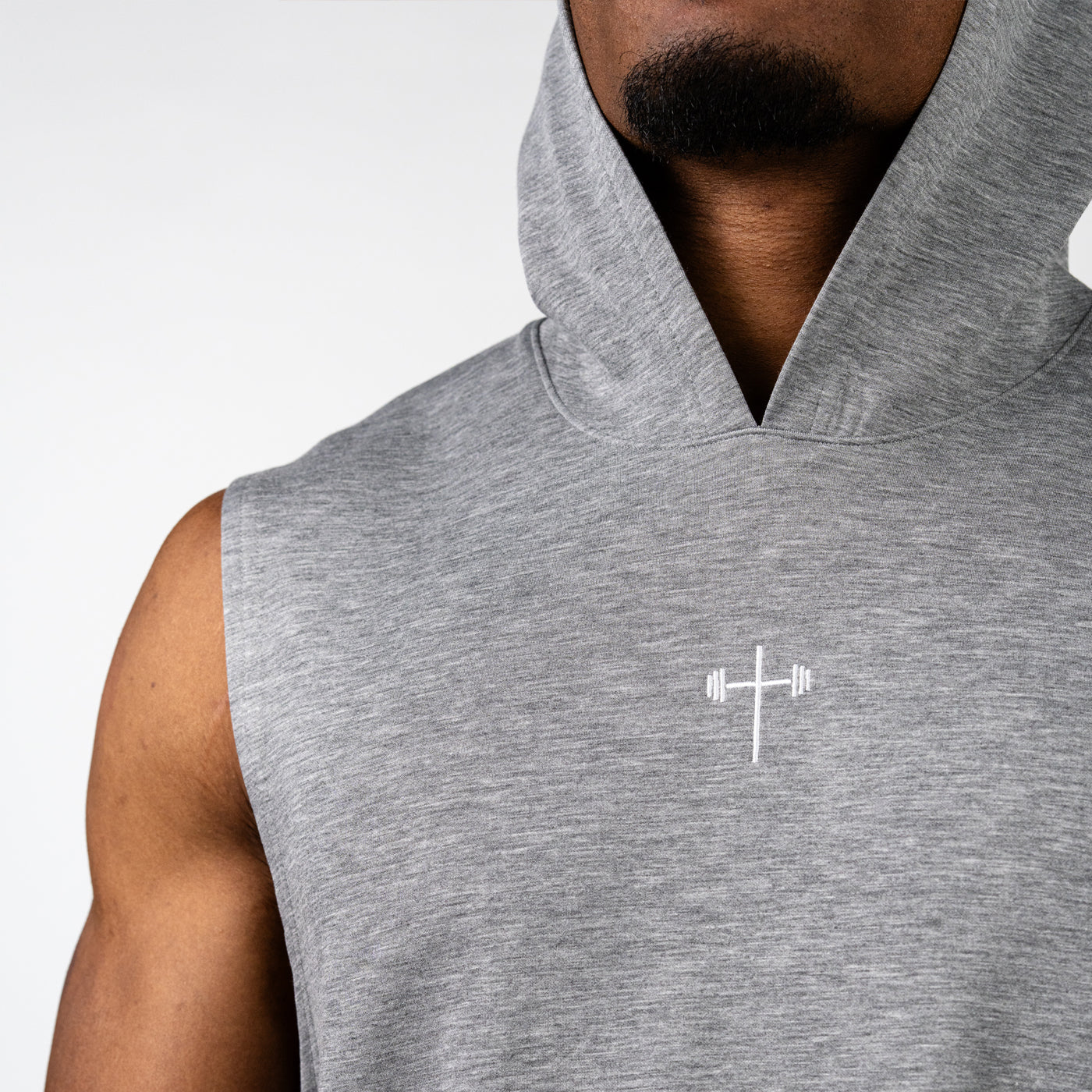 Close-up of a man wearing a sleeveless gray hoodie featuring a small white barbell cross logo on the chest, highlighting Christian fitness apparel.