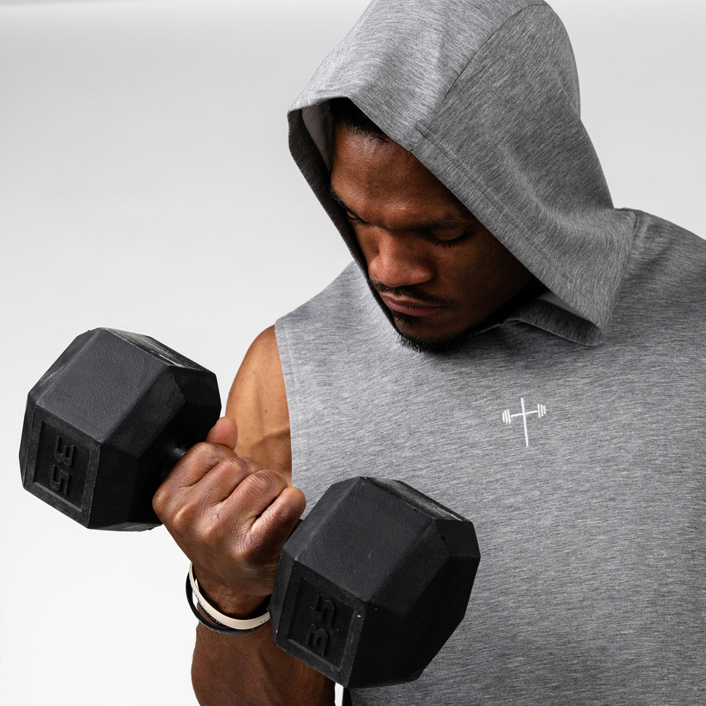 Man wearing a sleeveless gray hooded gym shirt with a small barbell cross logo lifting a black dumbbell during a workout session.