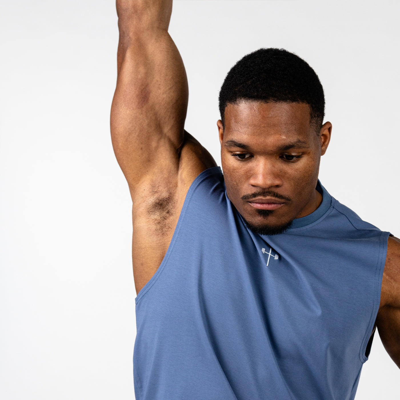 Athletic man wearing a sleeveless blue workout shirt with a small cross and barbell design on the chest, raising one arm against a plain white background.
