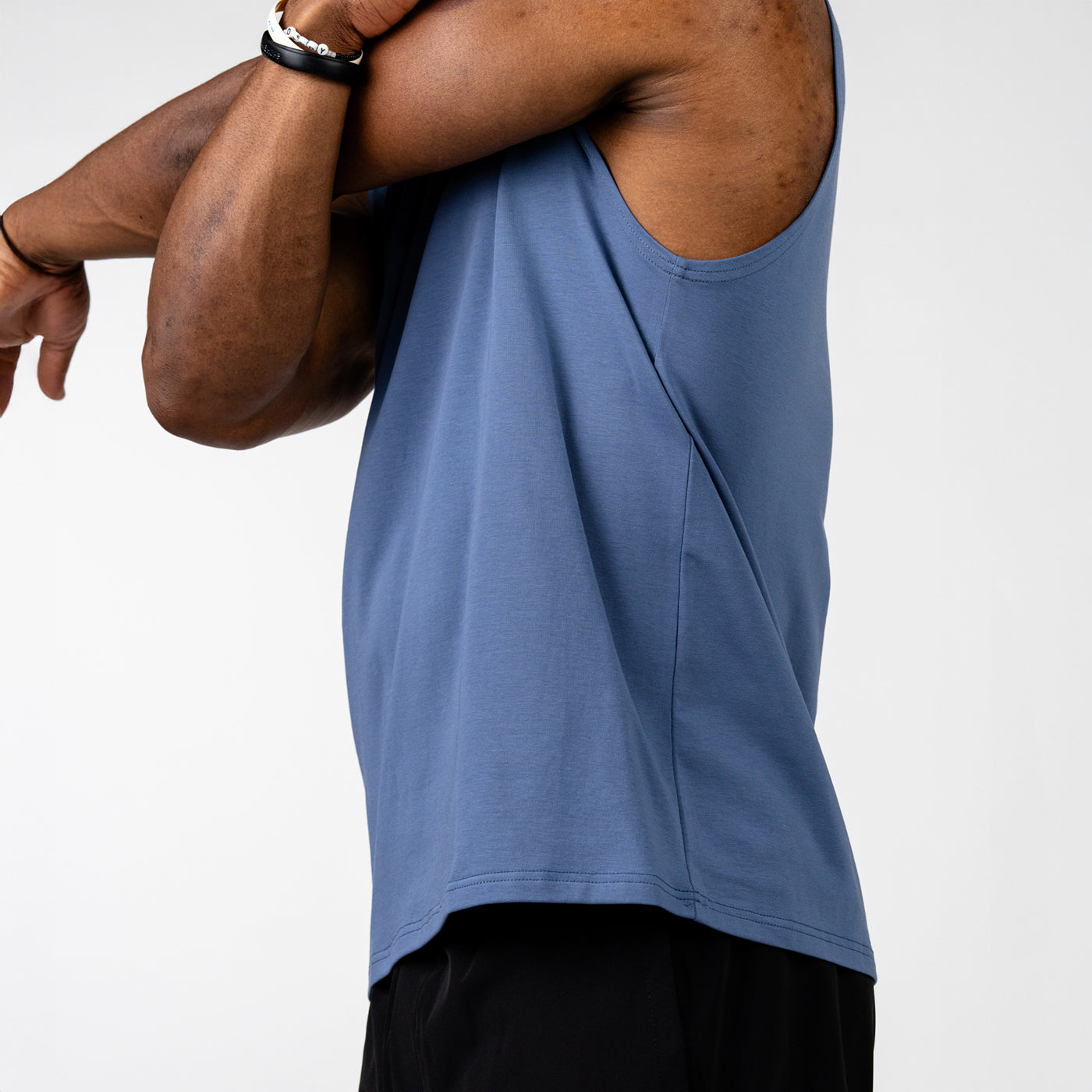 Close-up of a person stretching their arm across their chest wearing a sleeveless blue athletic shirt and black shorts against a plain white background.