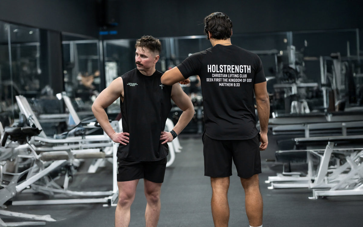 Two men wearing black sleeveless workout shirts and shorts stand in a gym, with one facing forward and the other showing the back of his shirt that reads "HOLSTRENGTH Christian Lifting Club" and a Bible verse reference, surrounded by gym equipment.