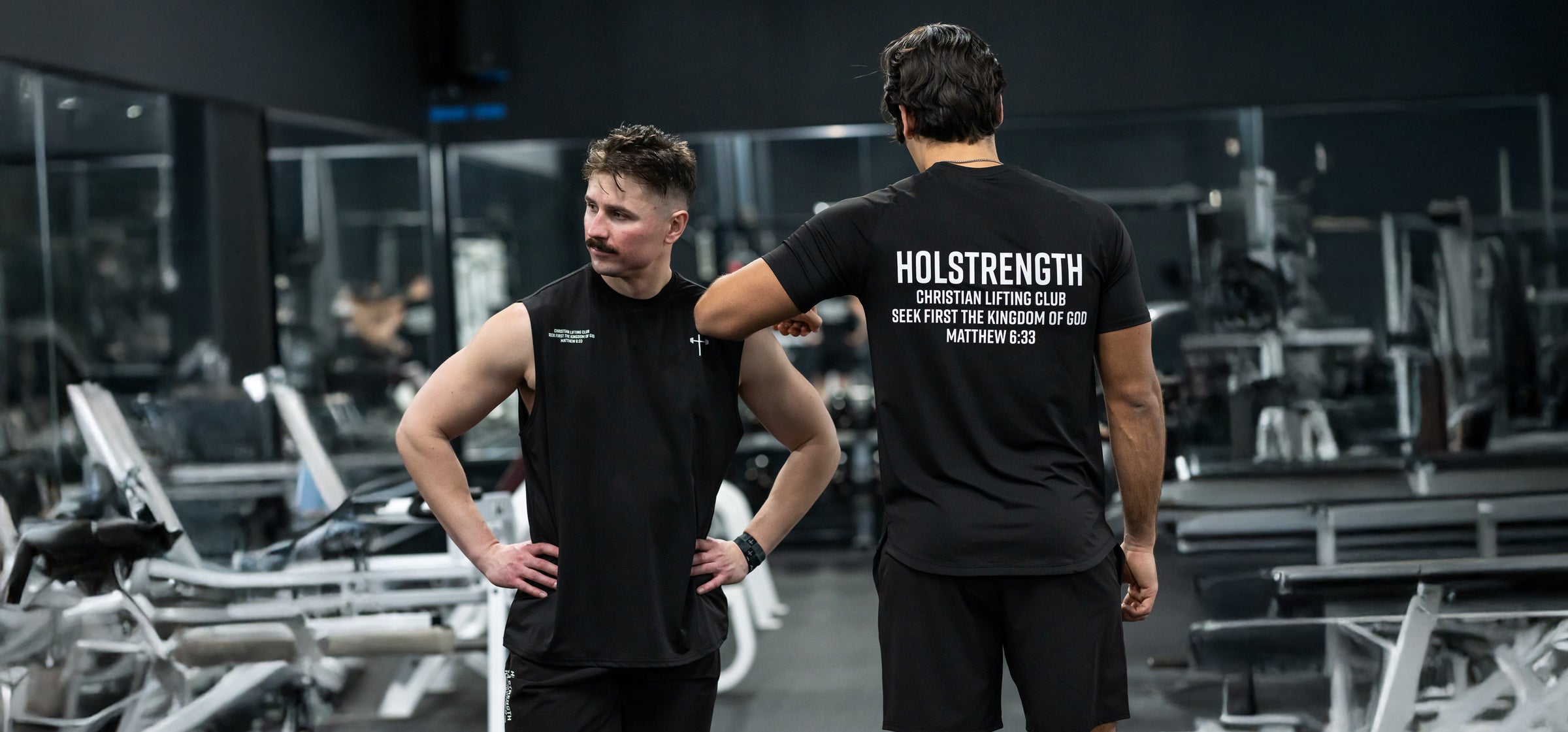 Two men in a gym setting wearing black sleeveless and short-sleeve shirts, one shirt displaying "HOLSTRENGTH Christian fitness club Seek first the kingdom of God Matthew 6:33" on the back.